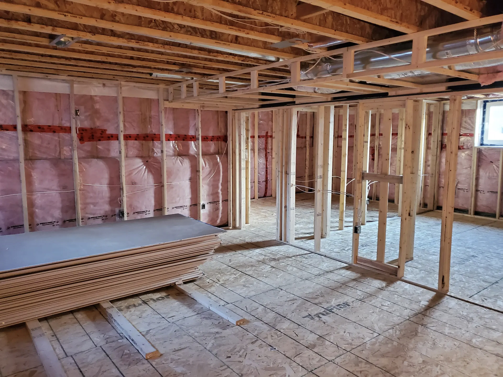 Basement under renovation with wood framing, installed insulation, visible ventilation ducts and stacks of drywall on the floor, ready for finishing work.