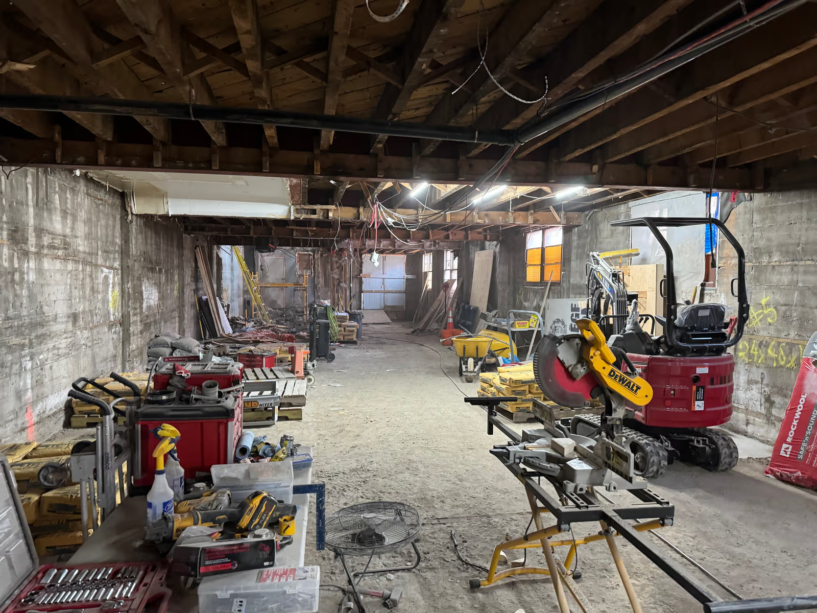 Chantier de rénovation intérieure avec structure de bois apparente, murs de béton, câblage visible, outils de construction, scie à onglets et mini-excavatrice 