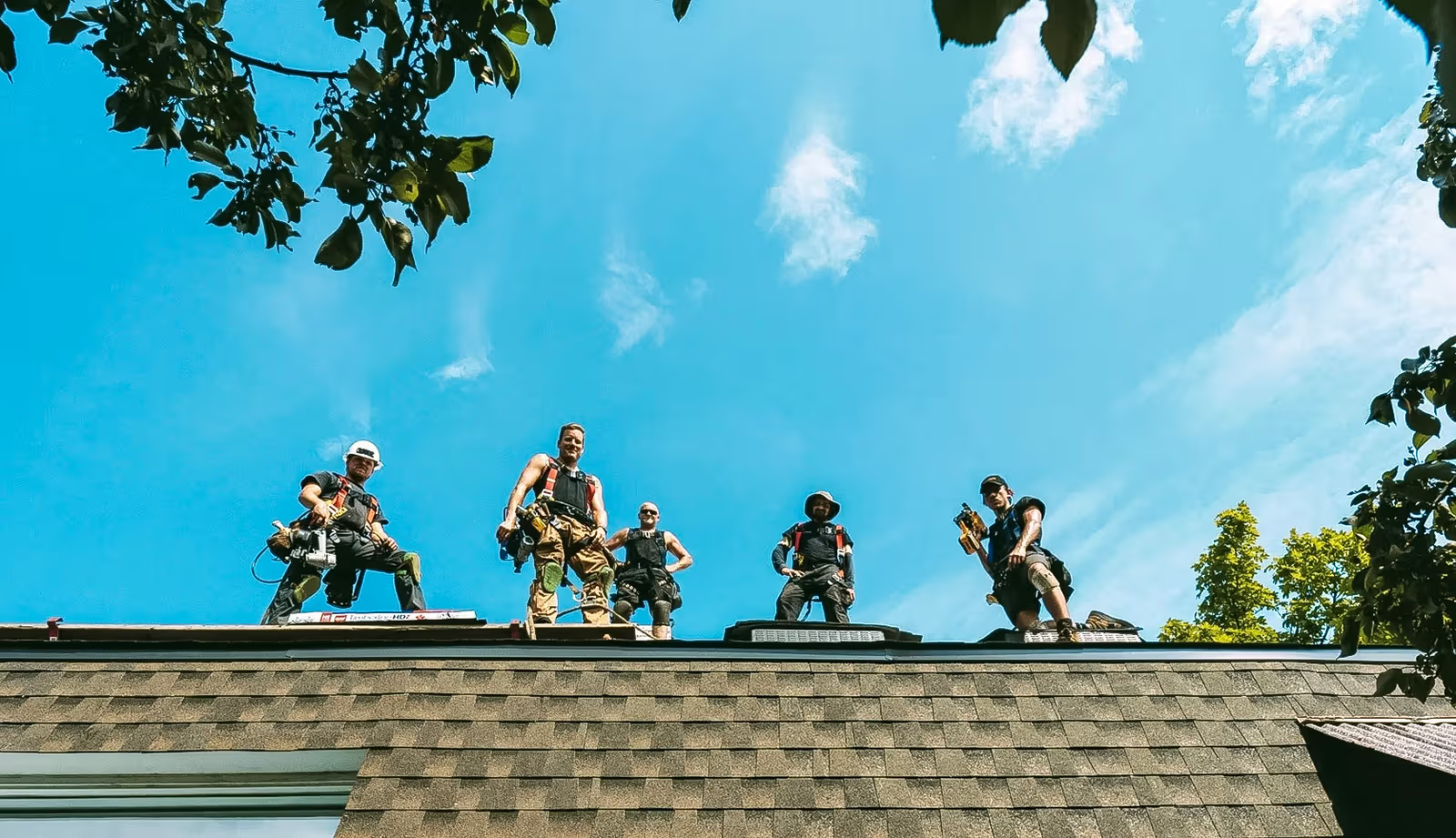 Équipe de couvreurs travaillant sur un toit en bardeaux sous un ciel bleu, avec harnais de sécurité, outils et arbres autour de la maison.
