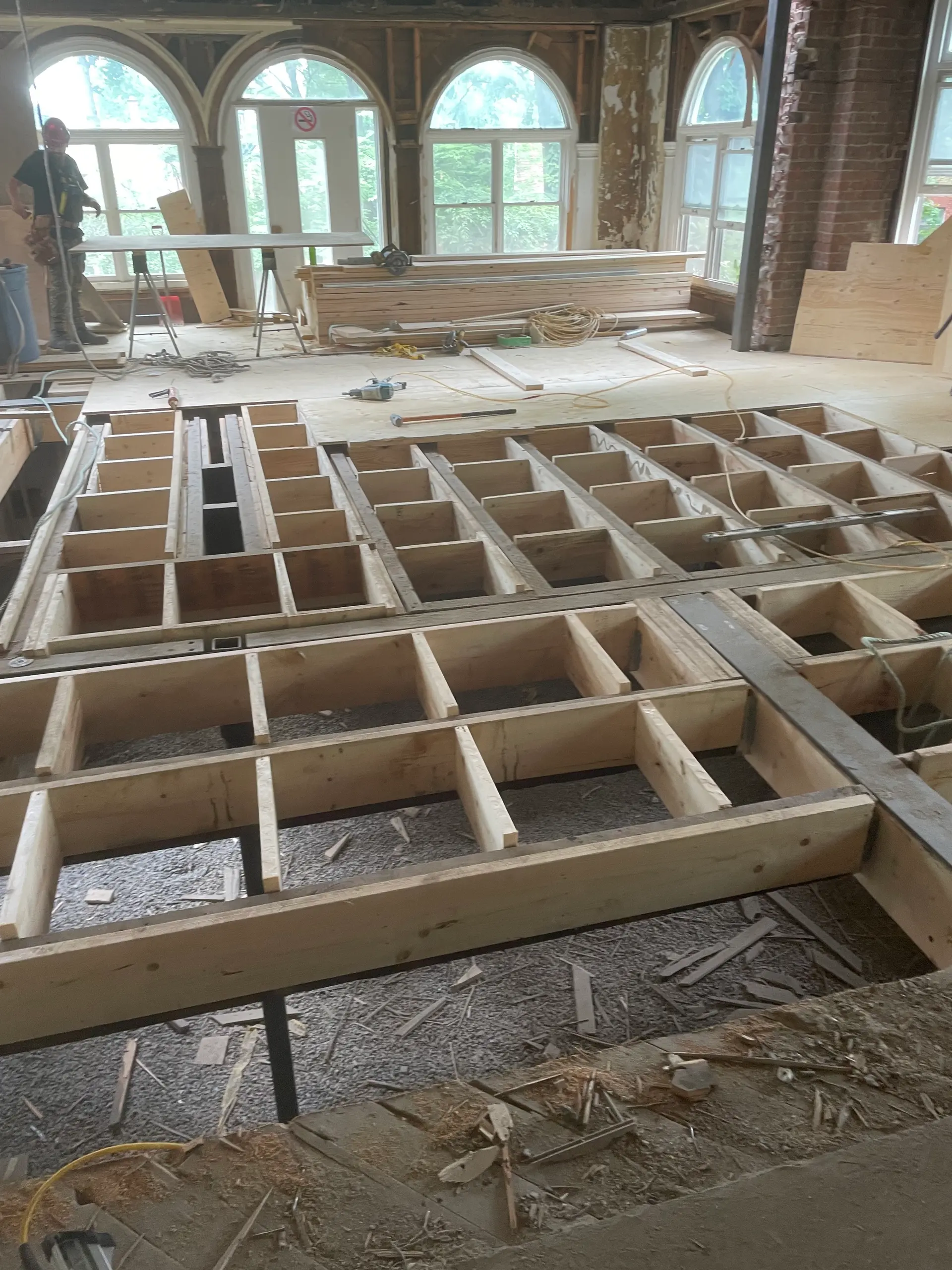 Interior reconstruction site with open floor framing, exposed joists, wood materials, and a worker on site.
