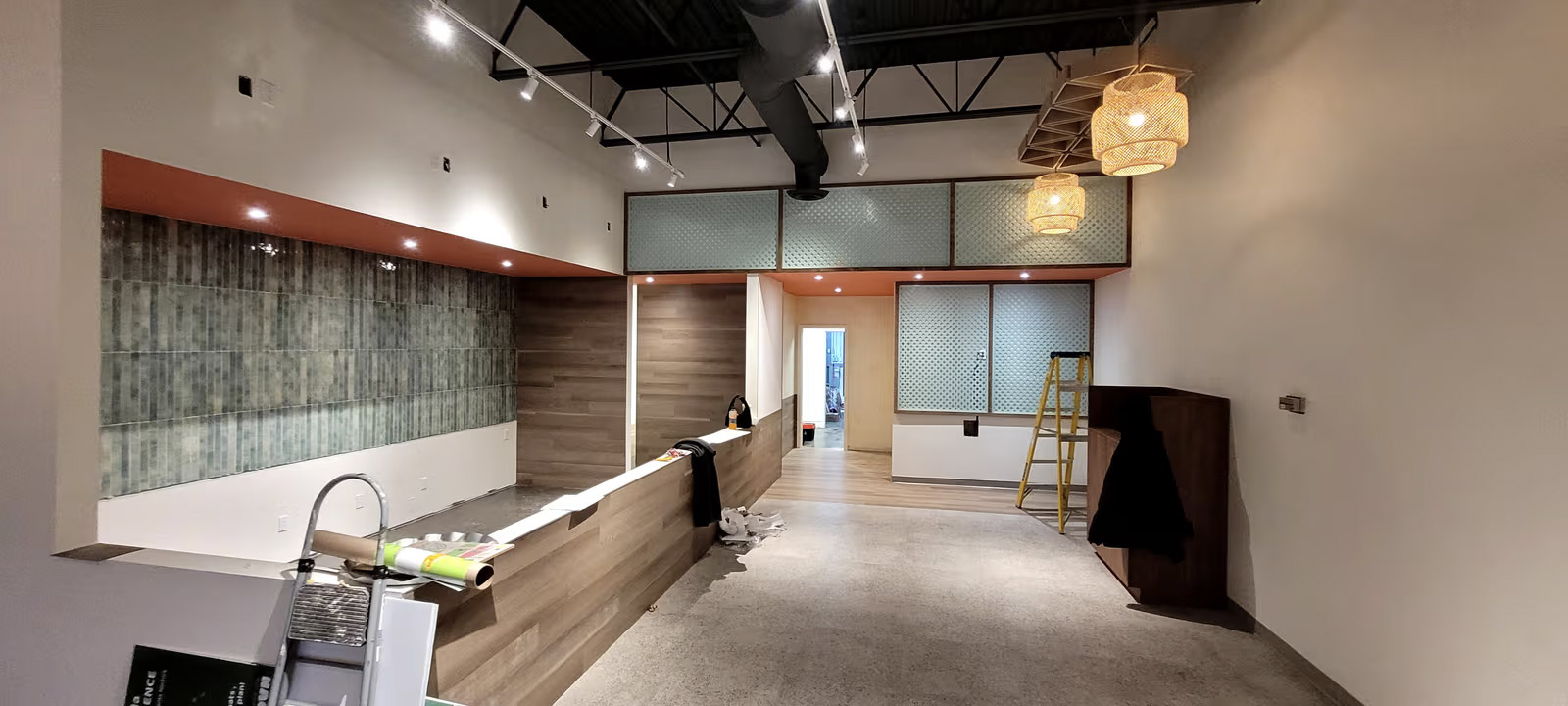 Local commercial en rénovation avec comptoir en bois, mur de céramique, éclairage sur rails, plafond industriel, conduits apparents et échelle de chantier.