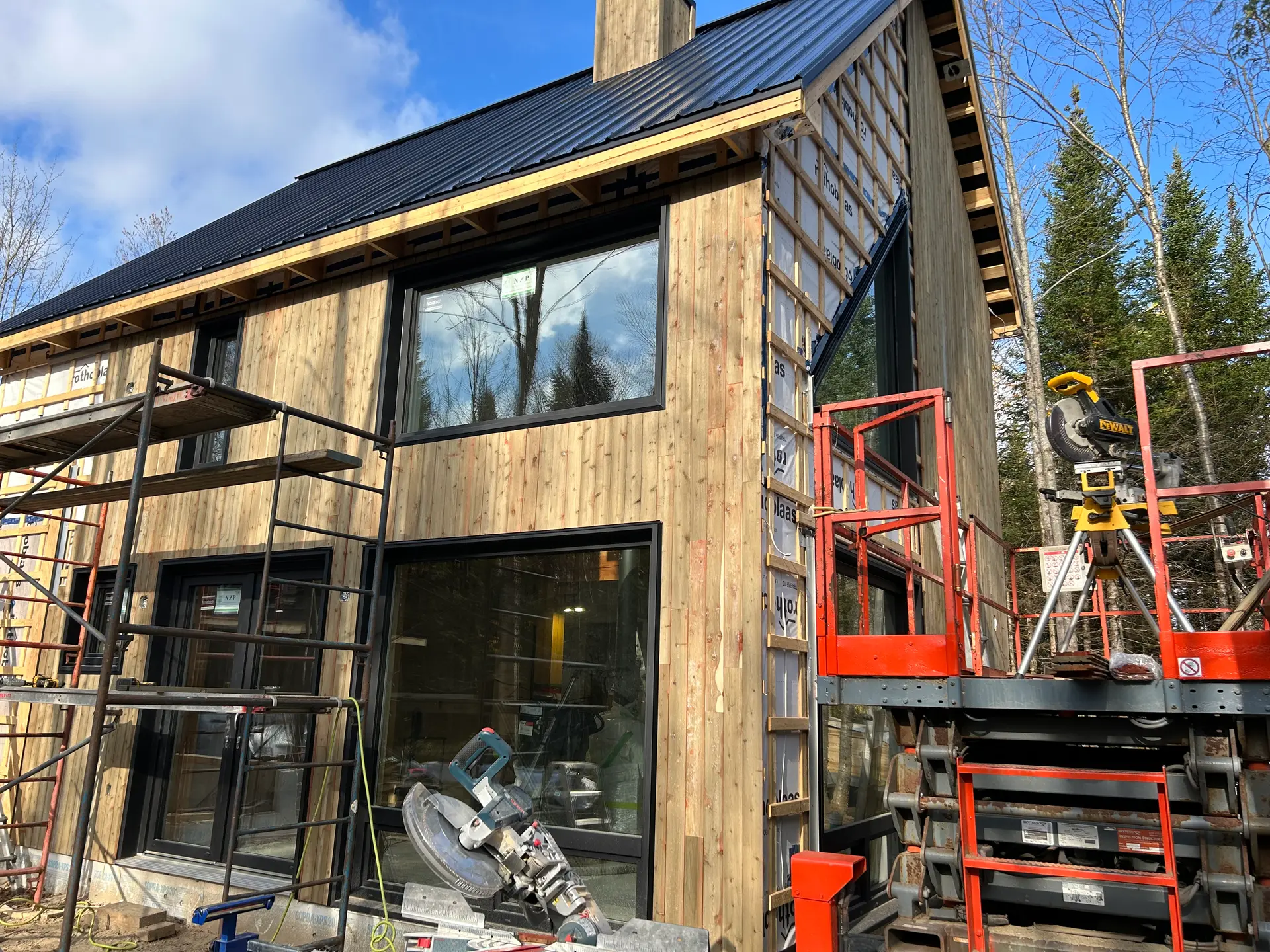 House under construction with wooden exterior cladding, large windows, and a black metal roof, surrounded by scaffolding and tools.