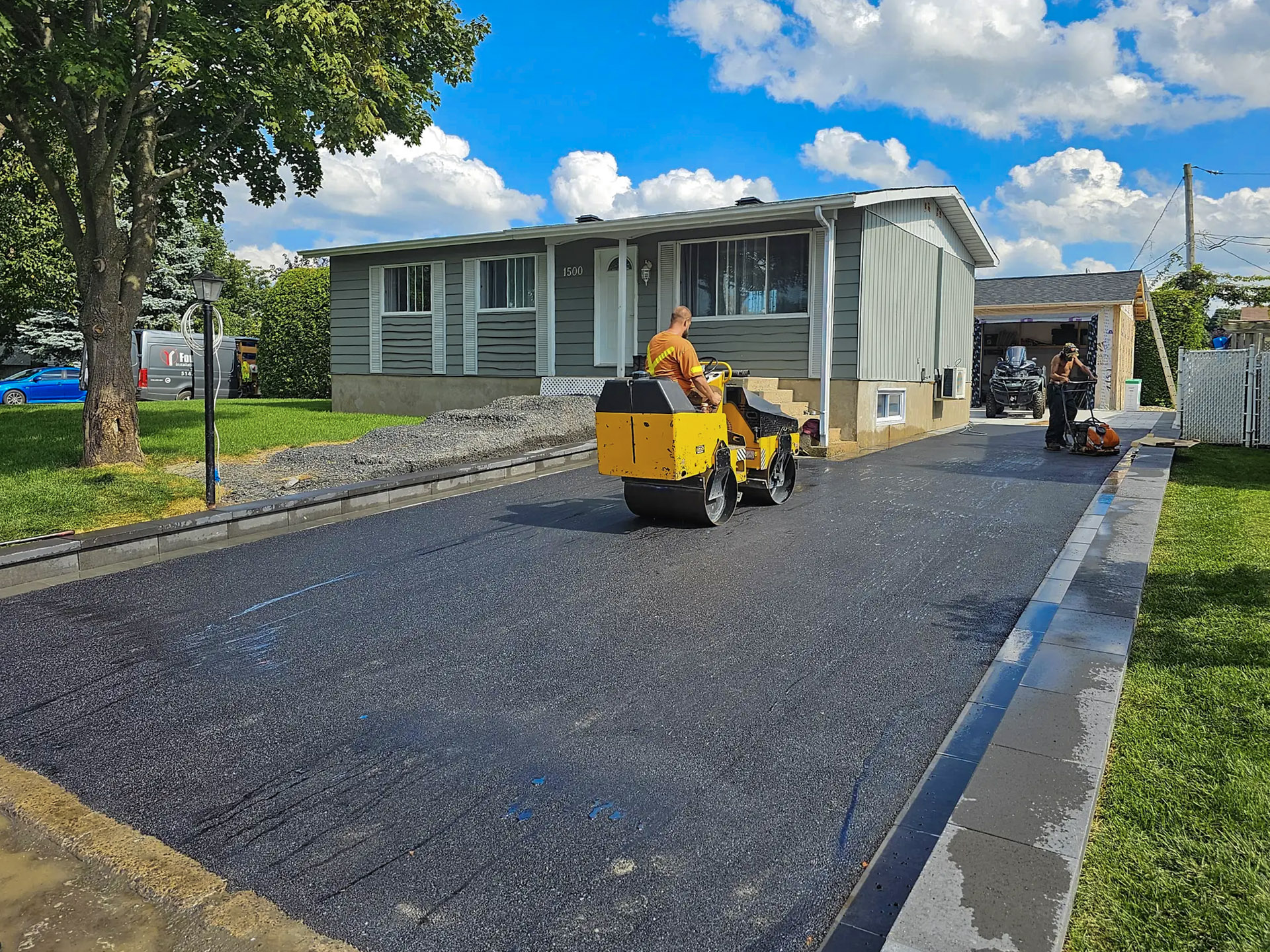 Travaux de pavage d'une allée résidentielle avec un rouleau compacteur et une plaque vibrante, devant une maison de plain-pied.