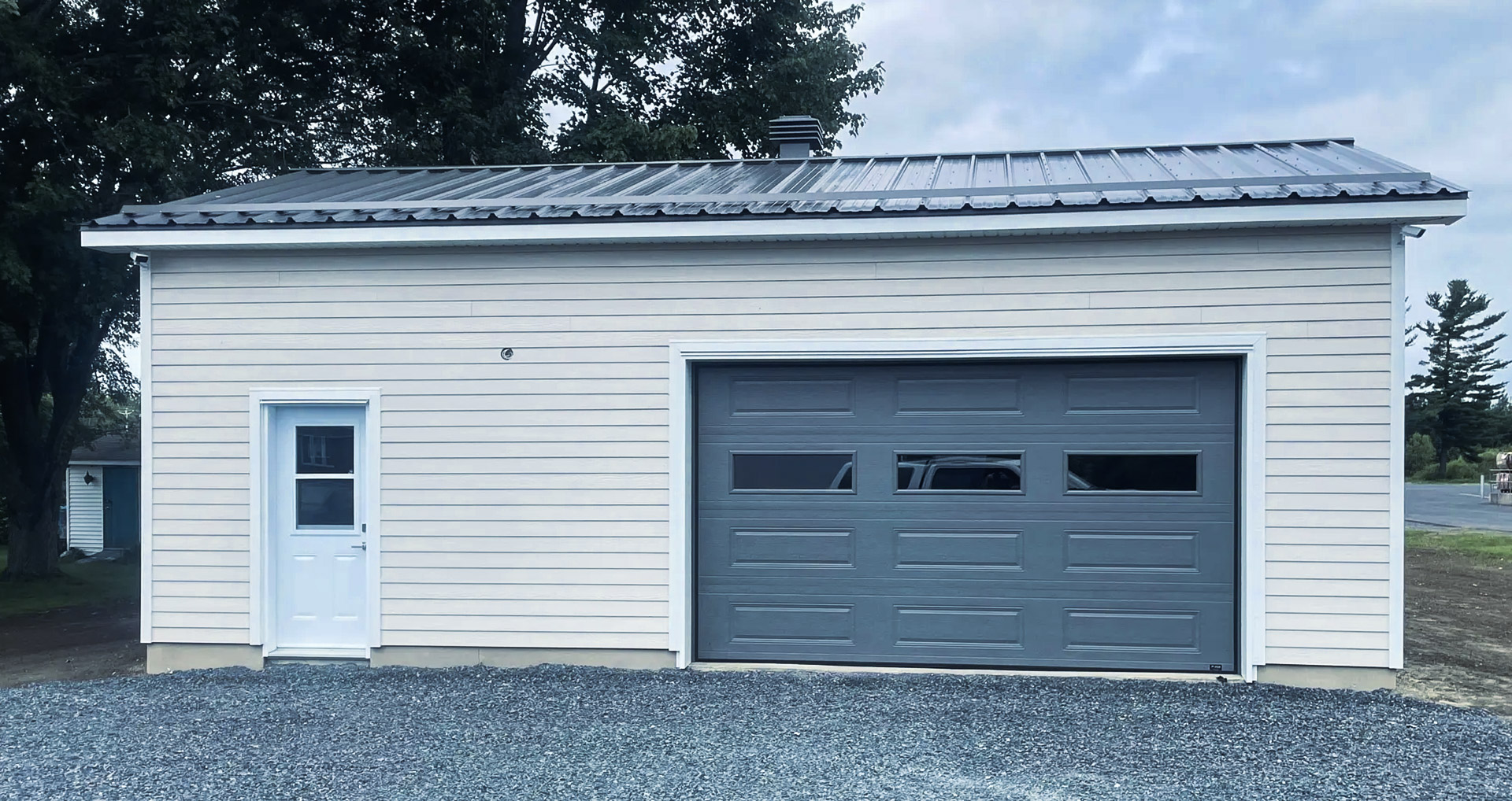 Garage moderne avec porte de garage sectionnelle grise et revêtement extérieur beige, toiture en tôle et entrée piétonne latérale sur sol en gravier.