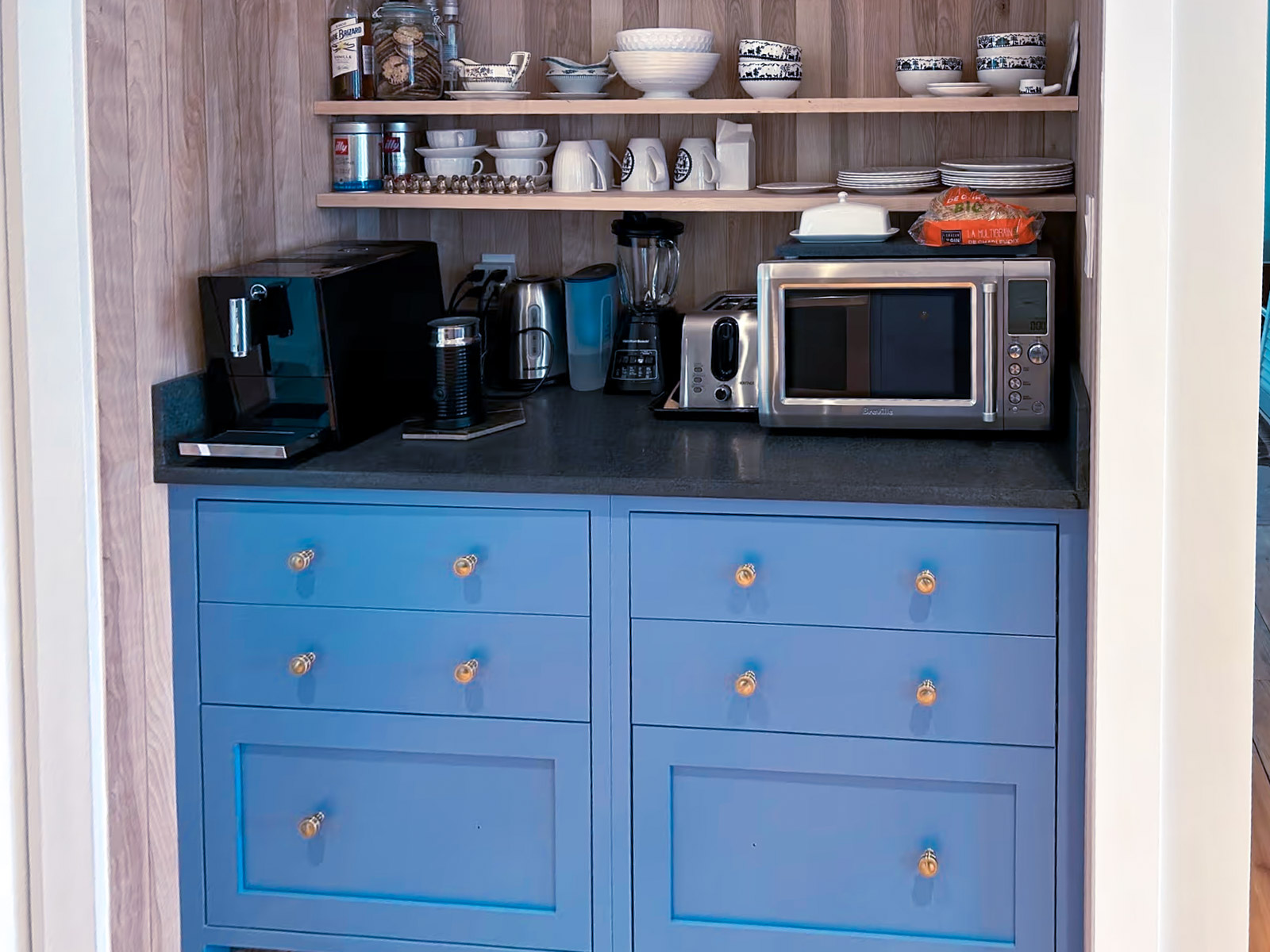 Built-in coffee station with custom blue cabinetry, black countertop, open wood shelving and small appliances in a modern kitchen