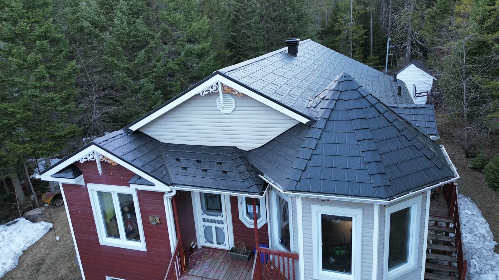 Forest home with a dark metal roof, red and grey siding, dormers, snow guards, and a wooden porch.
