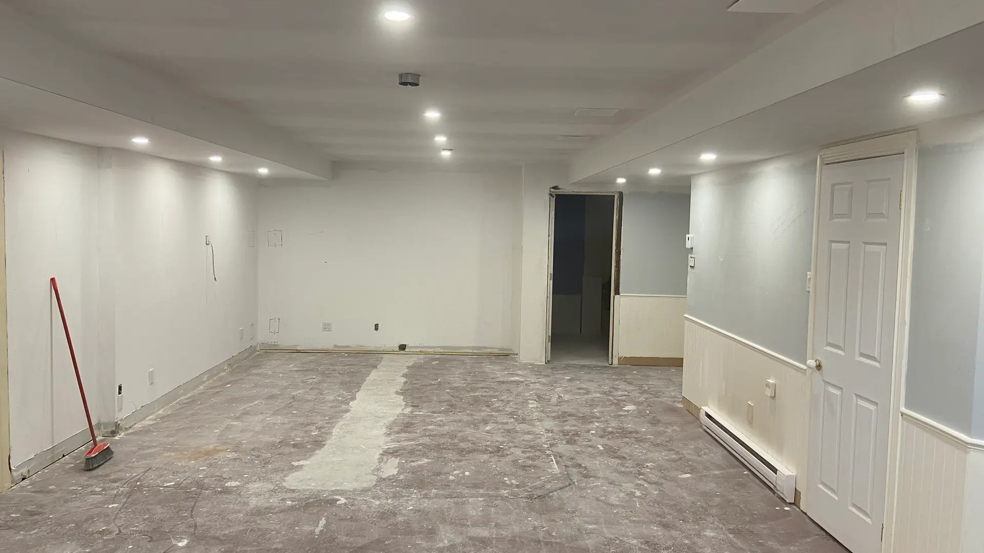 Sous-sol rénové avec murs peints, éclairage encastré, plancher retiré et balai de chantier.