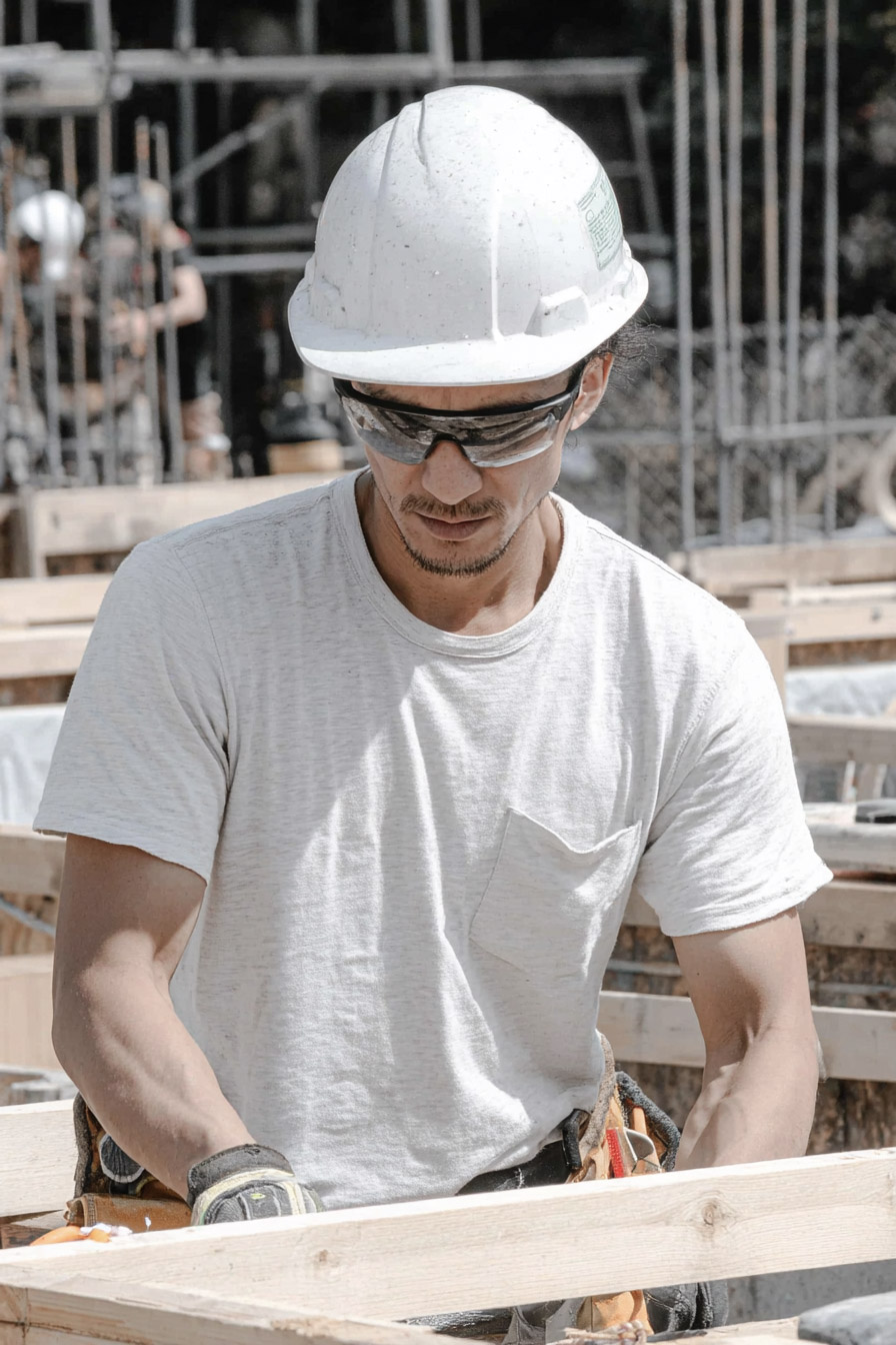 Ouvrier de construction avec casque et lunettes manipulant une planche de bois sur un chantier extérieur