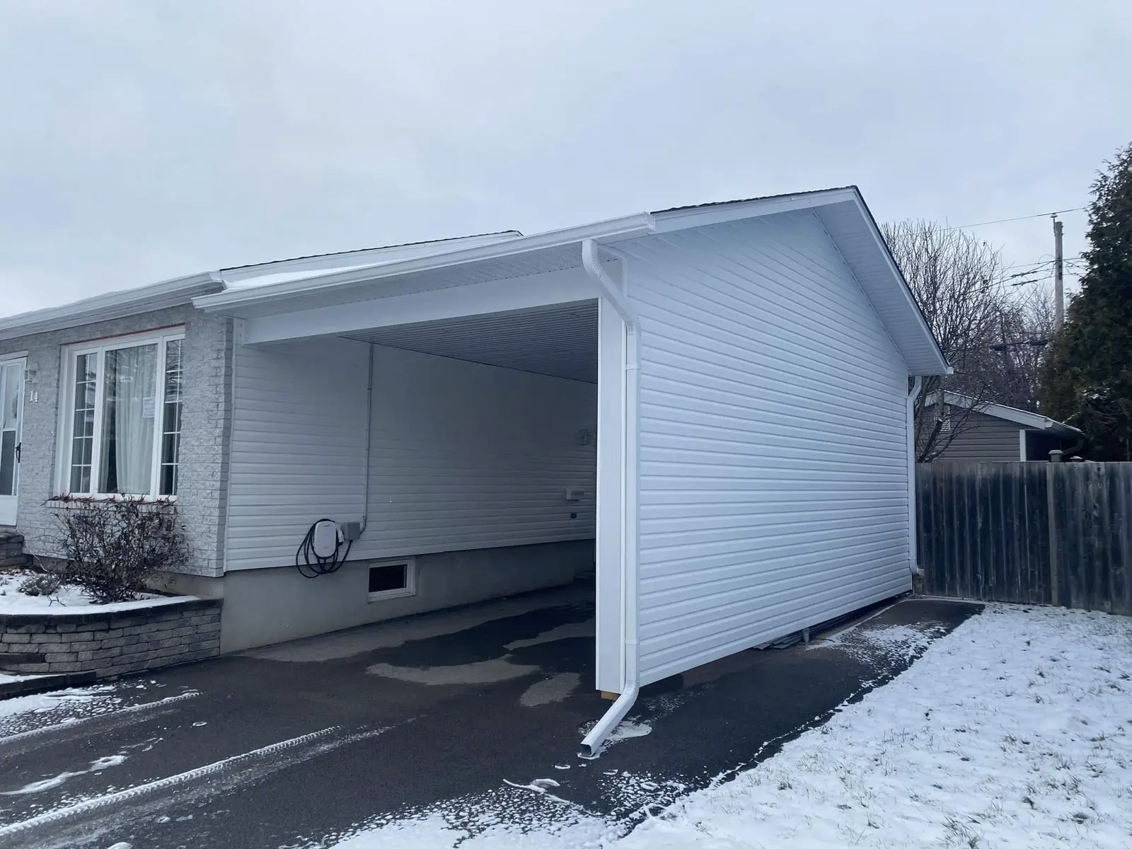 Abri d’auto blanc annexé à une maison avec revêtement extérieur, gouttières, soffites, entrée asphaltée et terrain enneigé.