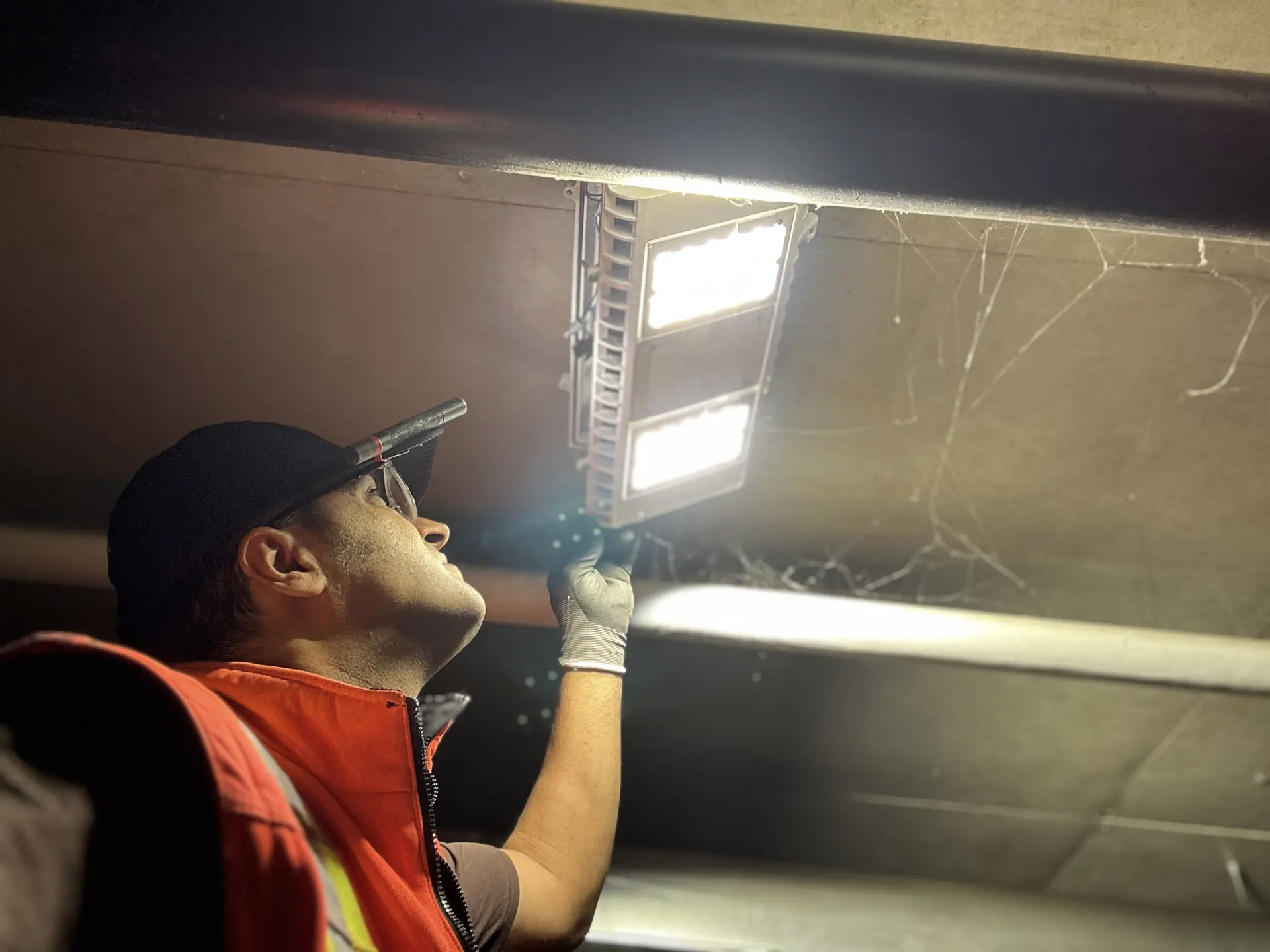 Électricien inspectant ou réparant un luminaire LED au plafond dans un espace sombre avec lampe frontale et gants de sécurité