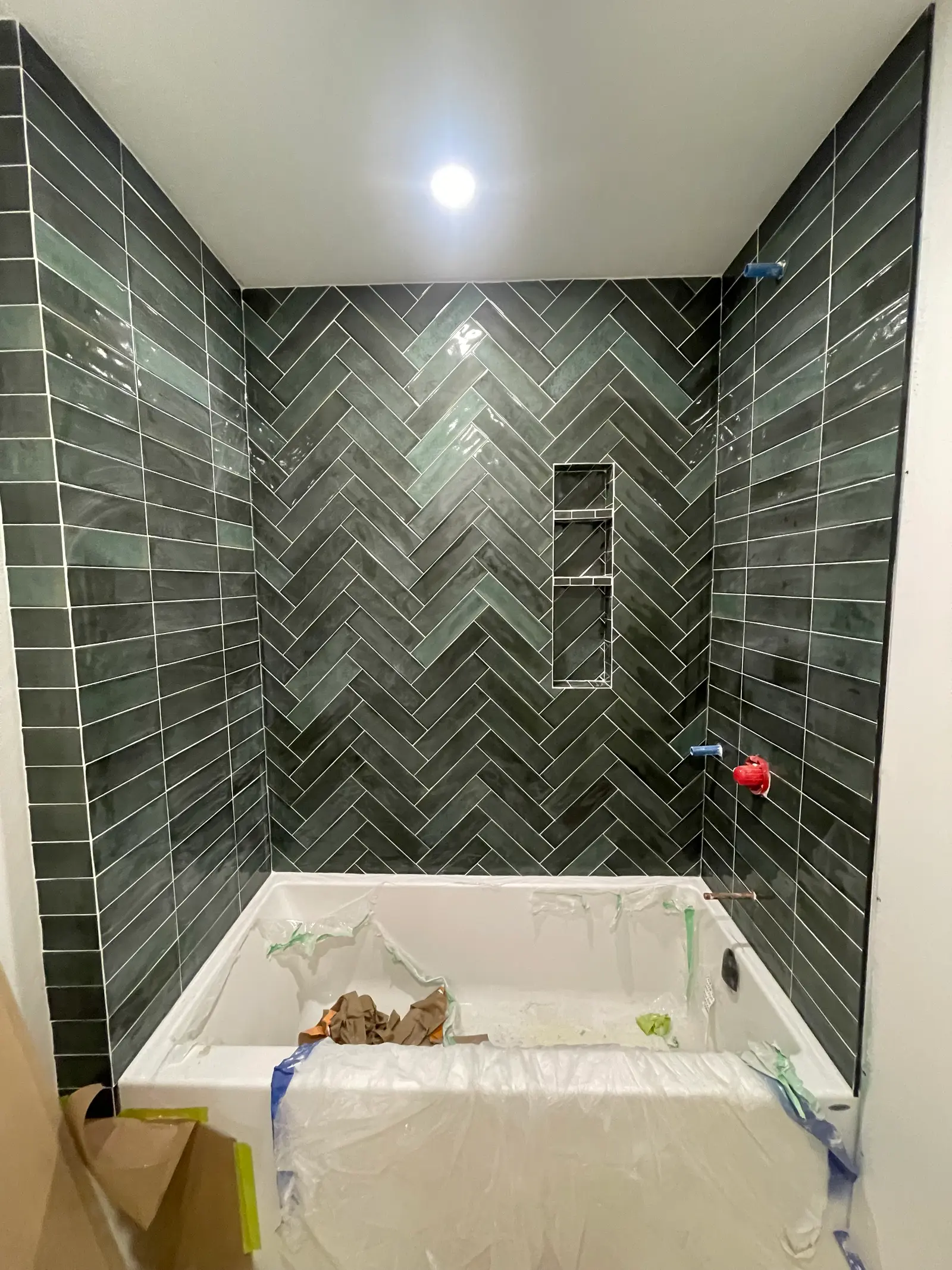 Salle de bain en rénovation avec baignoire blanche protégée, murs recouverts de carreaux vert foncé et motif chevron au fond de la douche.