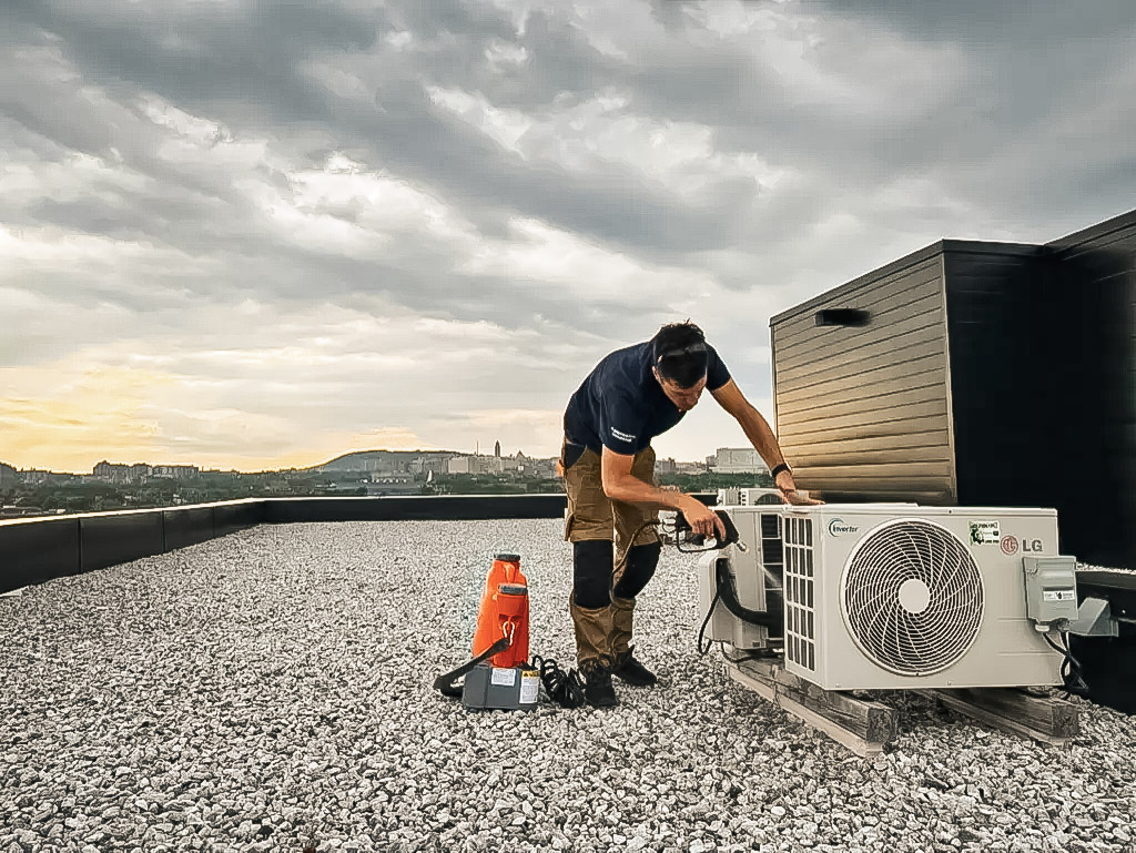 Technicien effectuant l’installation ou la maintenance d’une unité extérieure LG sur le toit en gravier d’un bâtiment, avec vue sur la ville en arrière-plan.