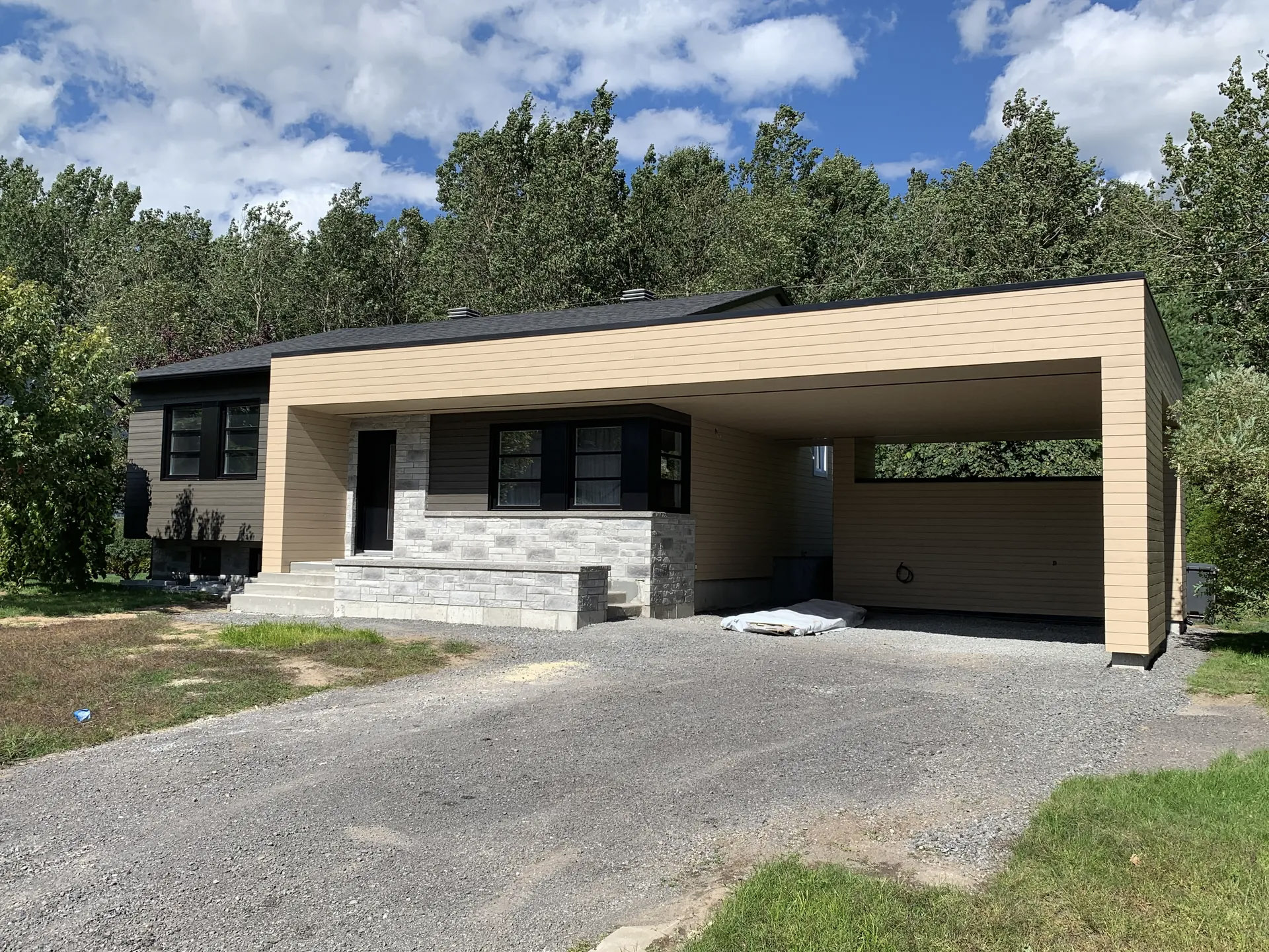 Maison moderne avec abri d’auto intégré, revêtement extérieur beige, muret de pierre, entrée en gravier et toiture en bardeaux noirs.
