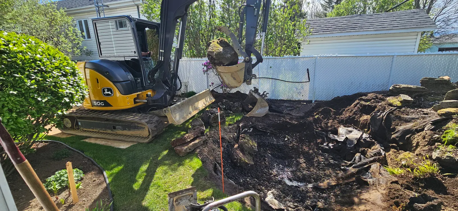 Excavator moving large rocks and soil during a backyard landscaping and drainage project.