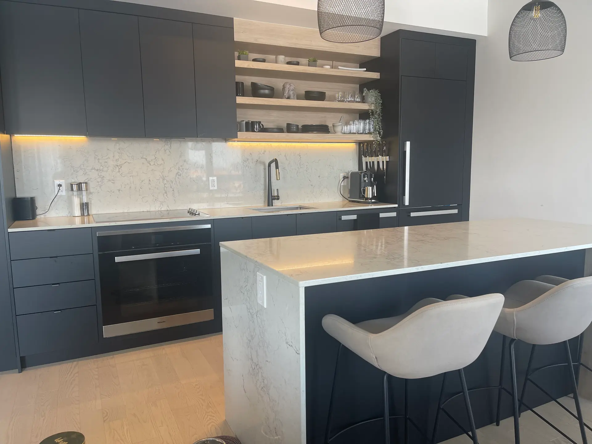 Cuisine moderne avec armoires noires mates, îlot en quartz blanc, dosseret marbré, tablettes en bois, luminaires suspendus et tabourets rembourrés