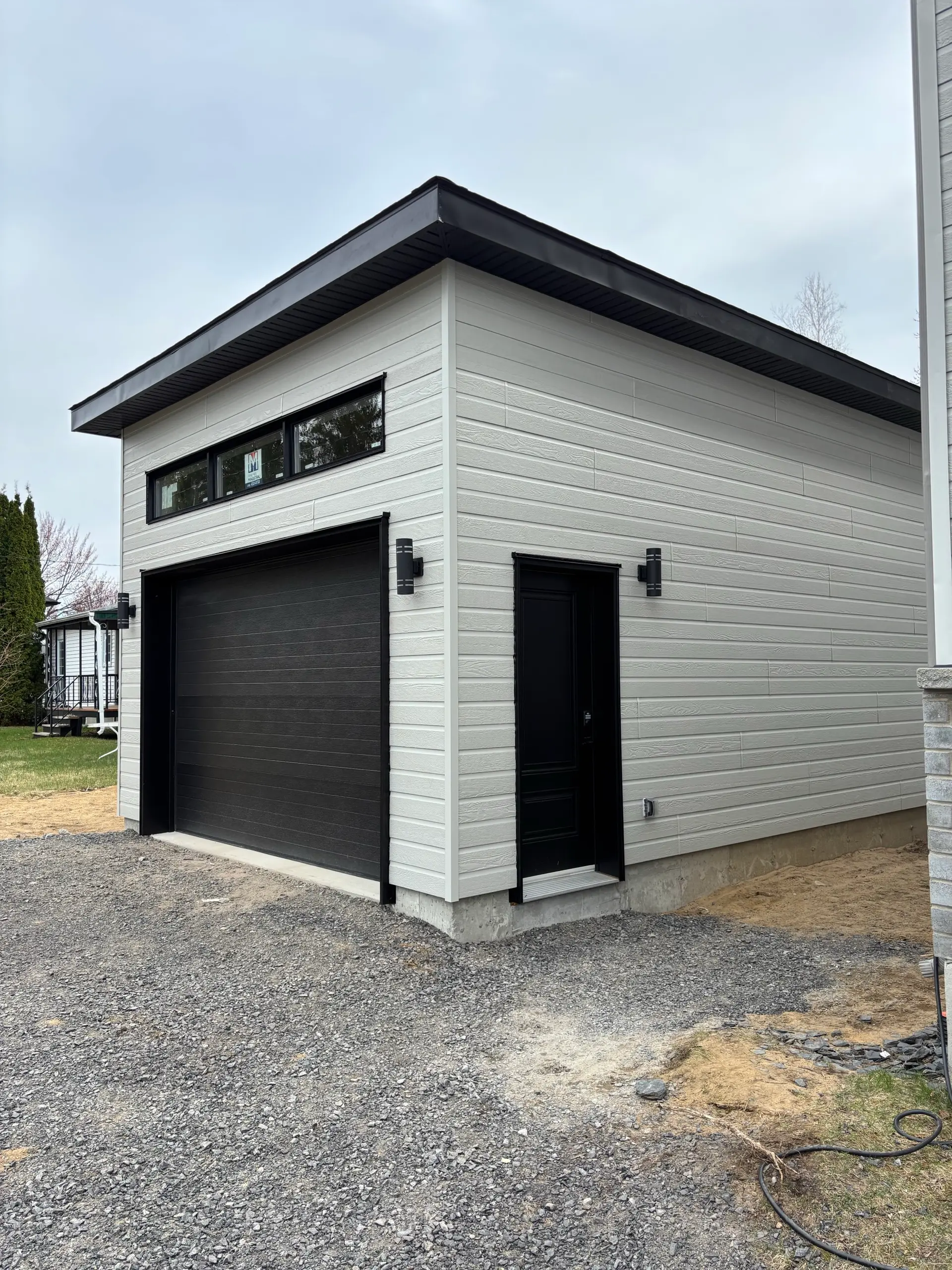 Modern detached garage with light gray siding, black door and side entrance on gravel lot