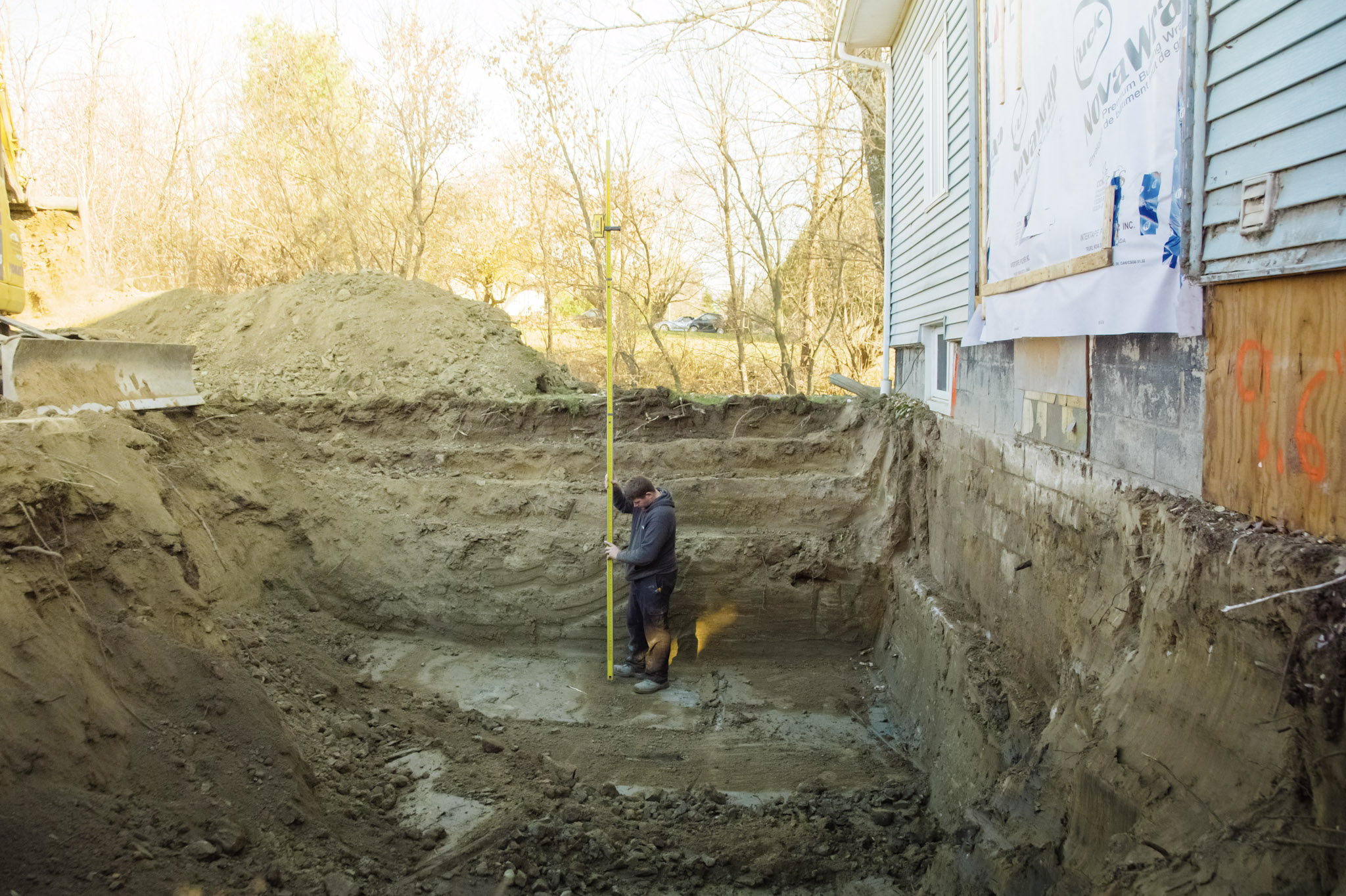 Worker measuring the depth of a foundation excavation using a grading rod near a house.