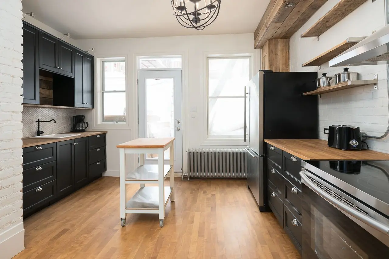 Bright kitchen with black cabinets, wood countertops, small white rolling island, wood flooring, white brick wall, open shelves and stainless steel appliances