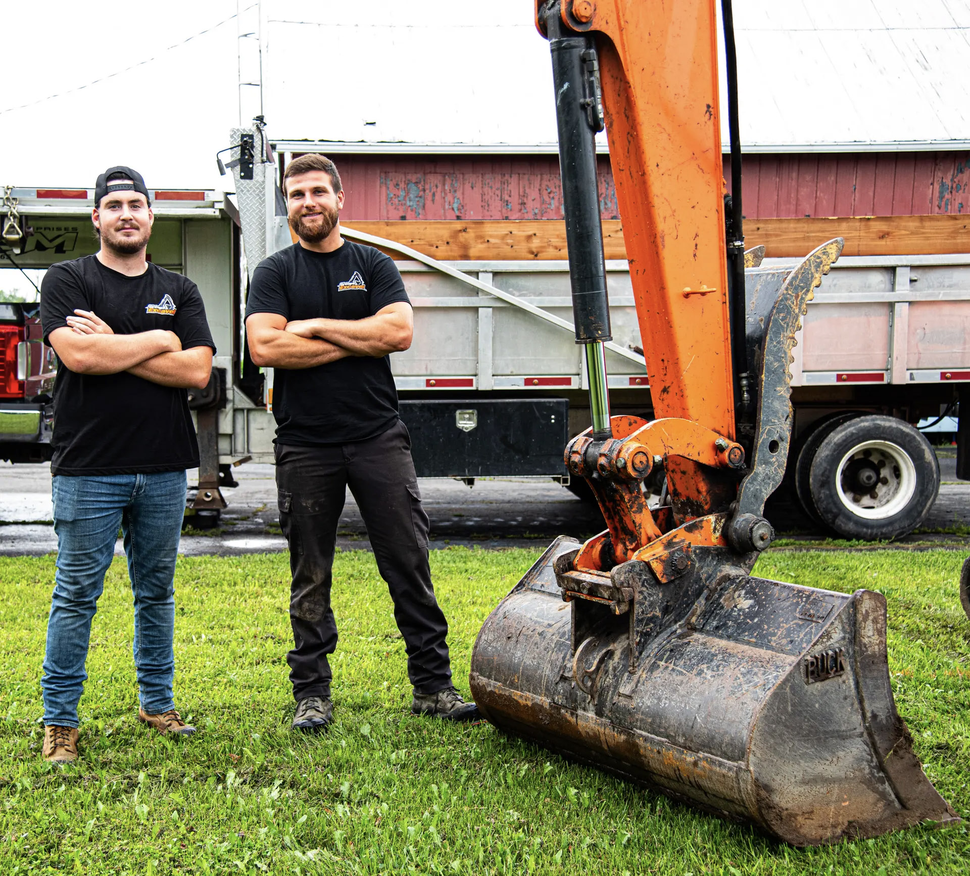 Two workers standing with arms crossed next to the bucket of an orange excavator, with a dump truck in the background.