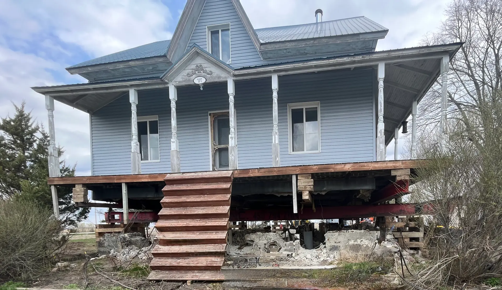 Old blue house raised for construction with wooden porch, white columns, metal roof and exposed foundation