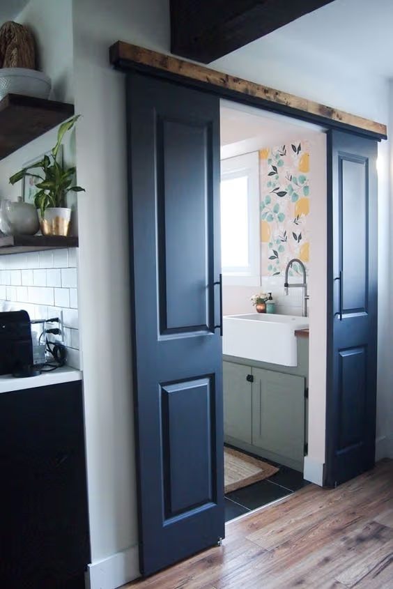 Portes coulissantes bleu foncé avec rail en bois et métal, ouvrant sur une petite salle de bain moderne avec lavabo blanc et papier peint floral.