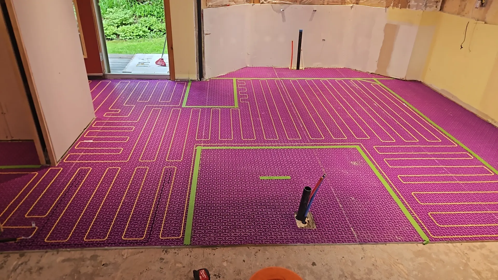 Installation de plancher chauffant hydronique avec membrane de désolidarisation mauve, tuyaux rouges, ruban vert et sorties de plomberie dans une pièce en rénovation