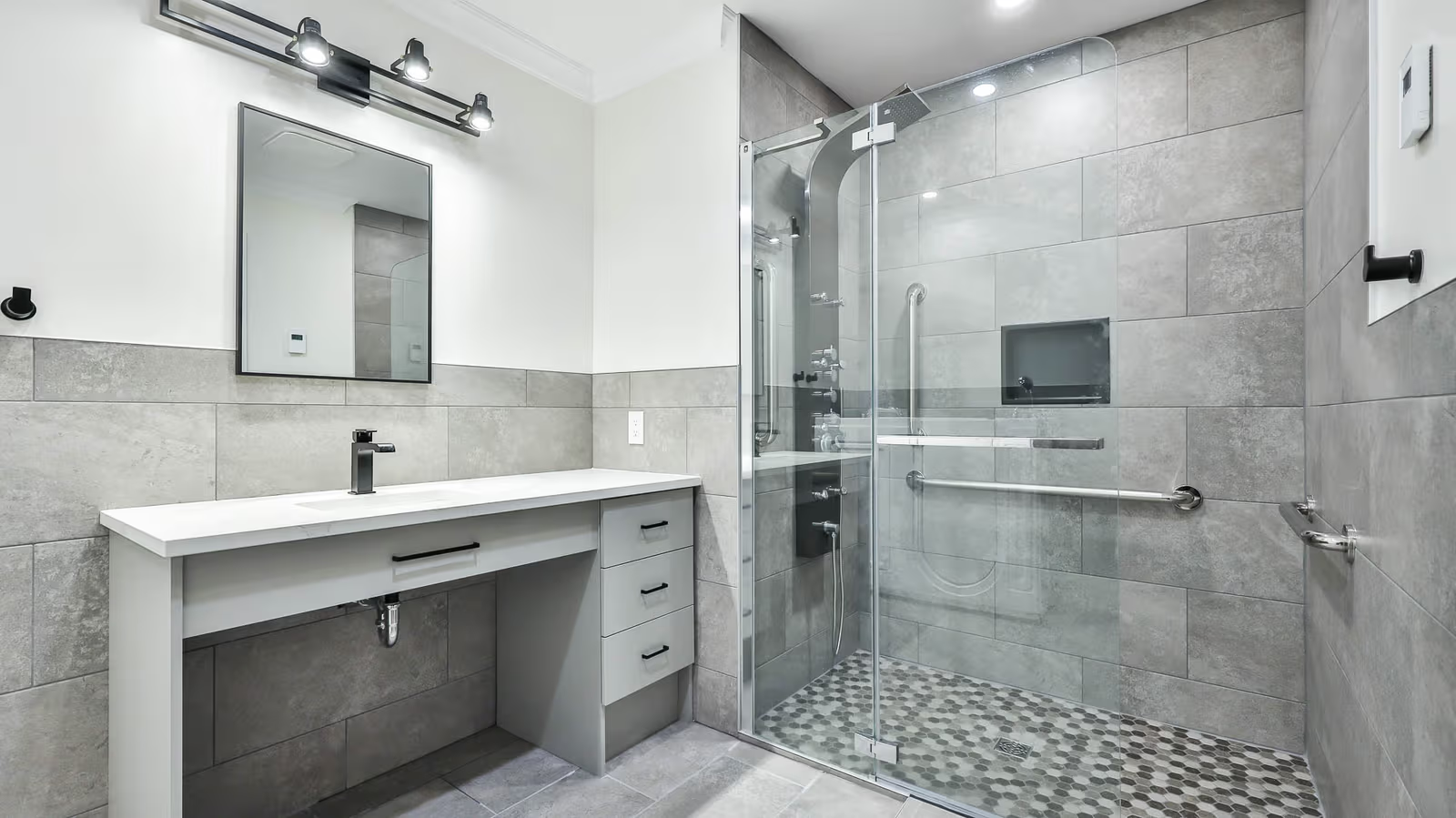 Salle de bain moderne avec douche vitrée accessible, carrelage gris, vanité blanche et robinetterie noire