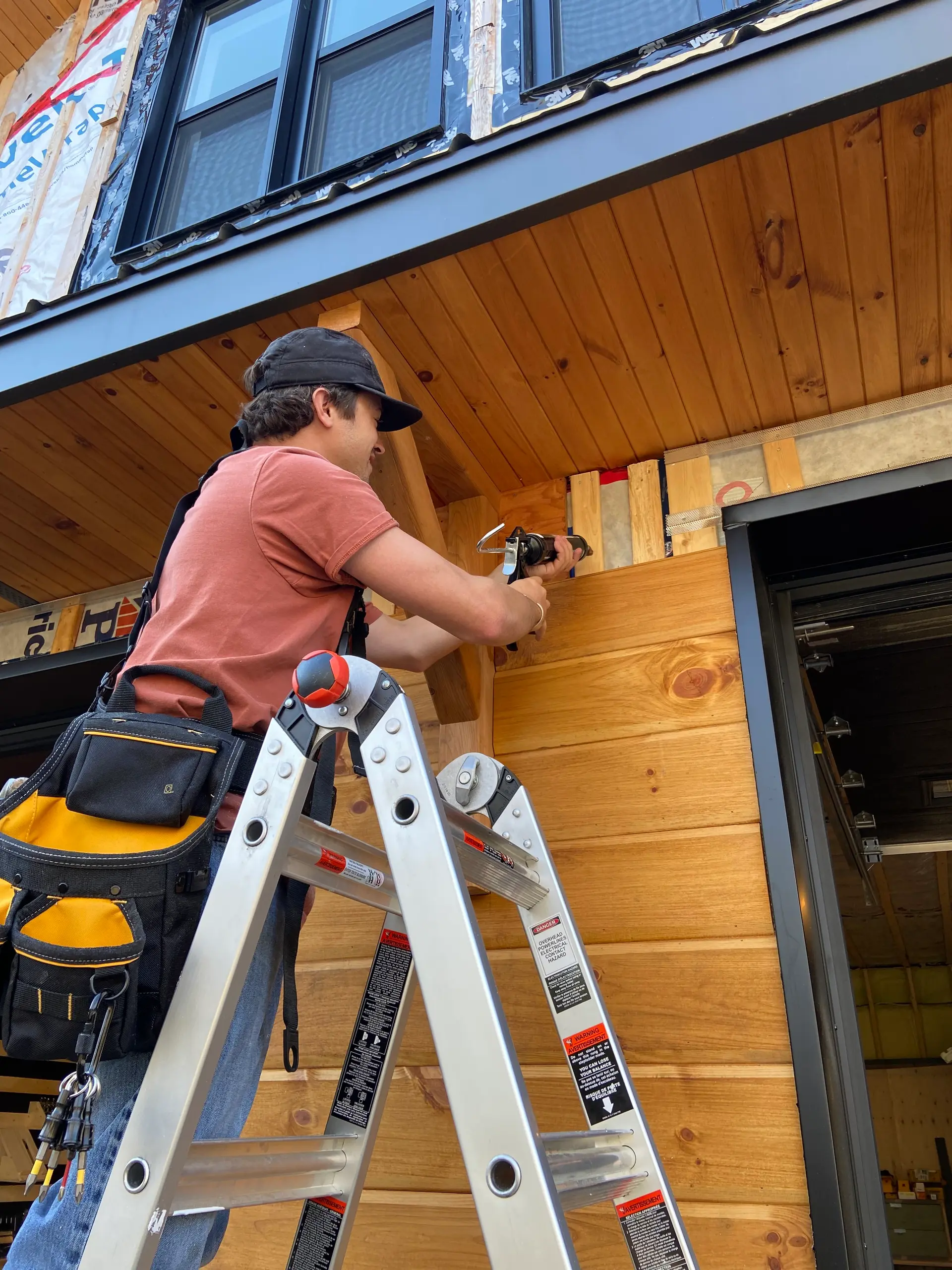 Travaux de rénovation avec installation de bardage en bois naturel par un ouvrier sur une échelle. /&nbsp;Renovation work with the installation of natural wood siding by a worker on a ladder.