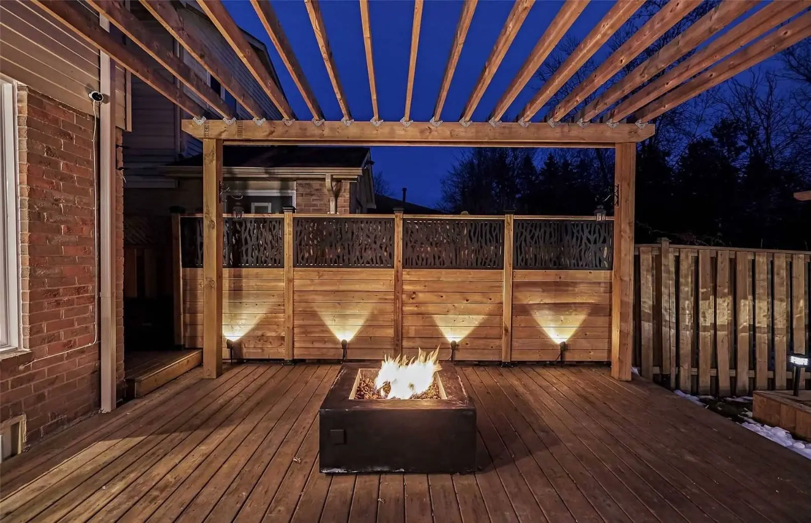 Terrasse en bois avec pergola et foyer extérieur illuminé, entourée d’une clôture en bois décorative, idéale pour des soirées chaleureuses en plein air.