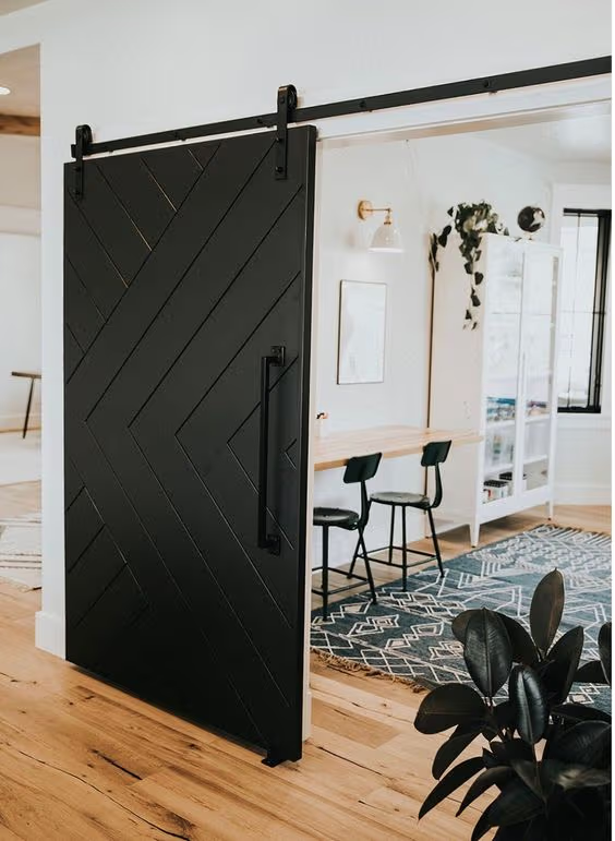 Porte coulissante noire à motif chevron avec rail apparent, séparant un espace bureau lumineux au style moderne et chaleureux.