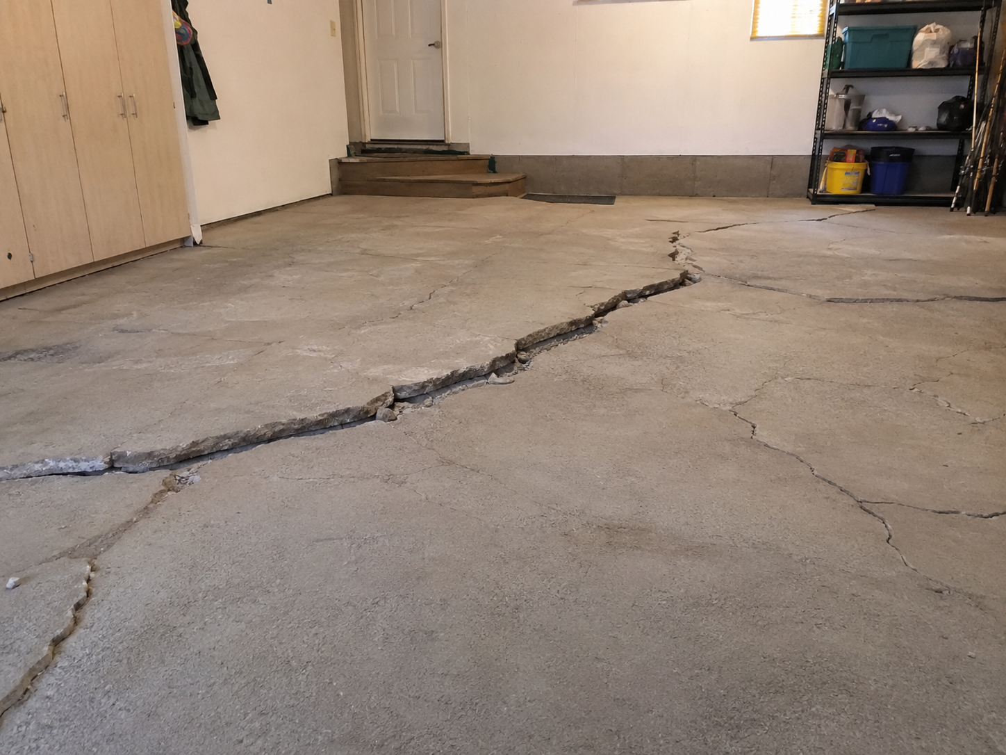 Un garage ou un sous-sol résidentiel avec de longues fissures irrégulières traversant le plancher de béton, signe d’un mouvement structurel.
