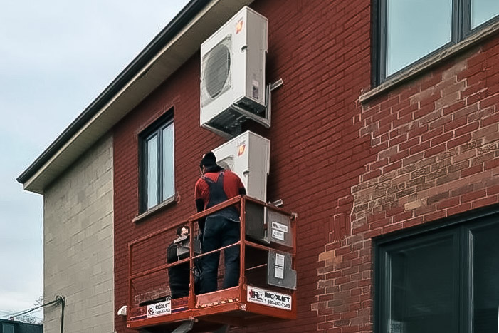 Deux techniciens sur une nacelle élévatrice installent des unités extérieures de climatisation sur un mur en brique.