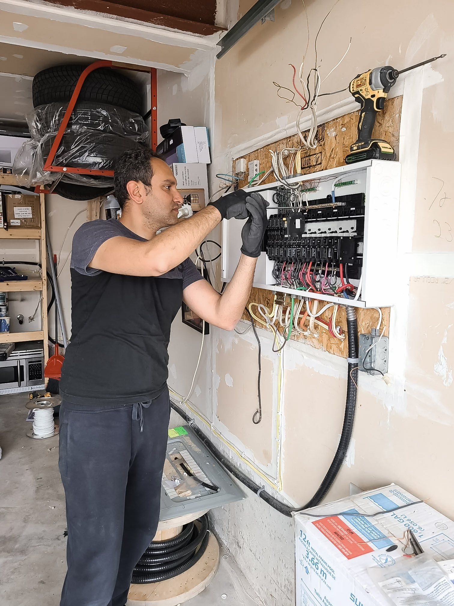 An electrician working on a circuit breaker panel in a garage, with exposed wiring and tools.