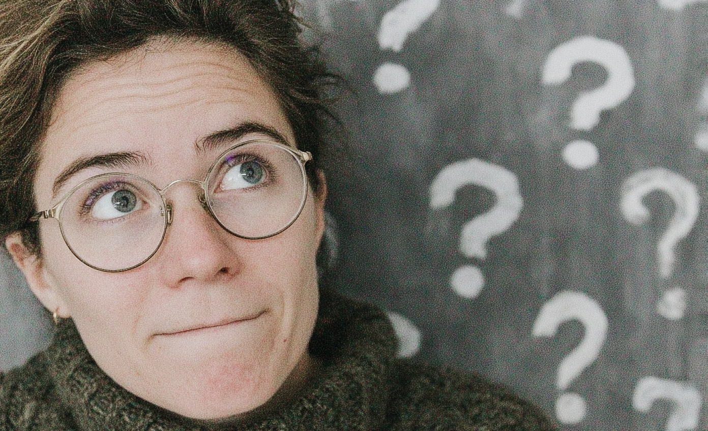 Personne pensive avec lunettes rondes regardant vers le haut, en arrière-plan un mur décoré de points d’interrogation blancs.