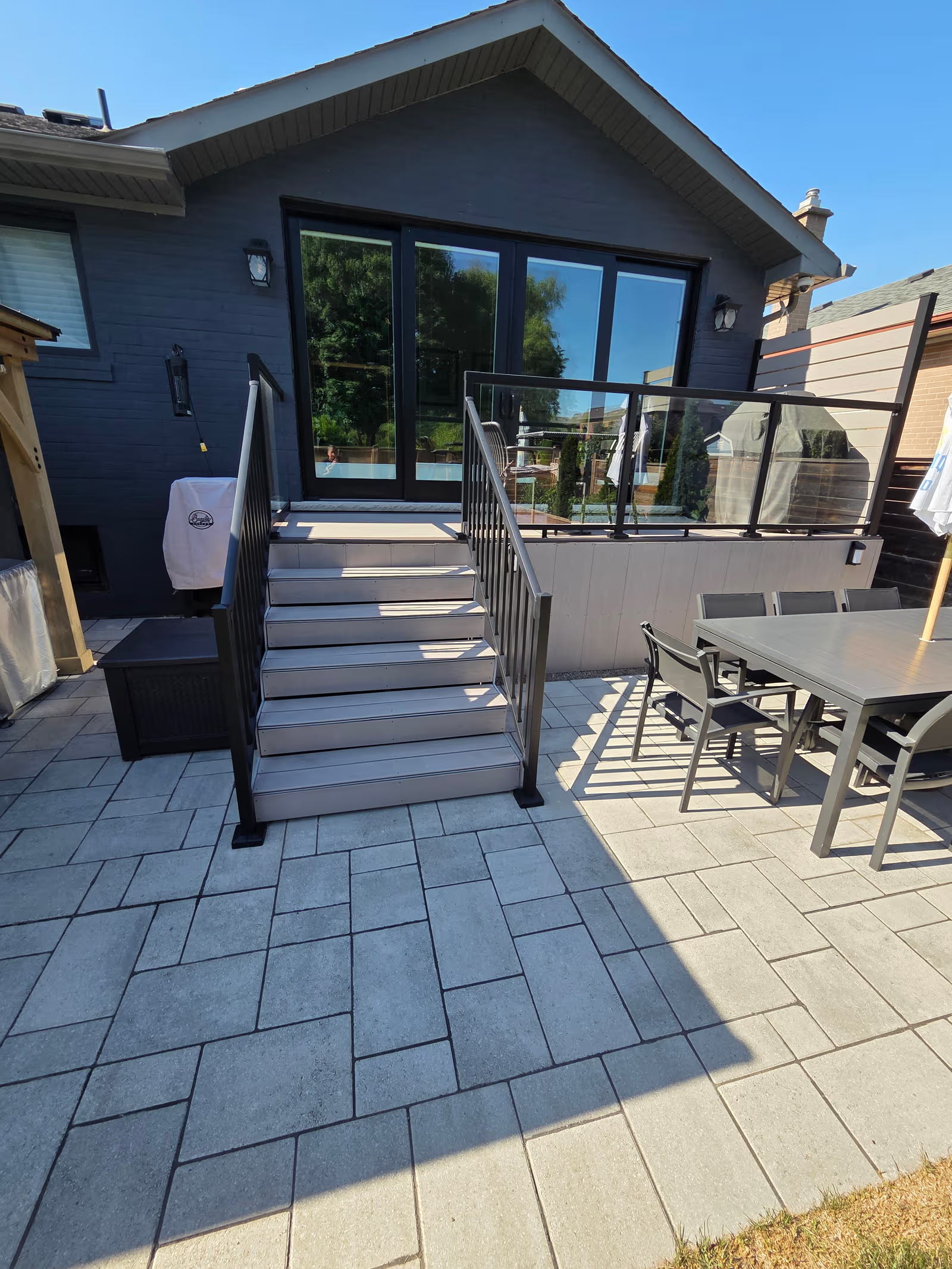 Terrasse moderne avec escalier en composite, rampes en verre et pavé uni, donnant accès à une maison avec grandes portes vitrées&nbsp;