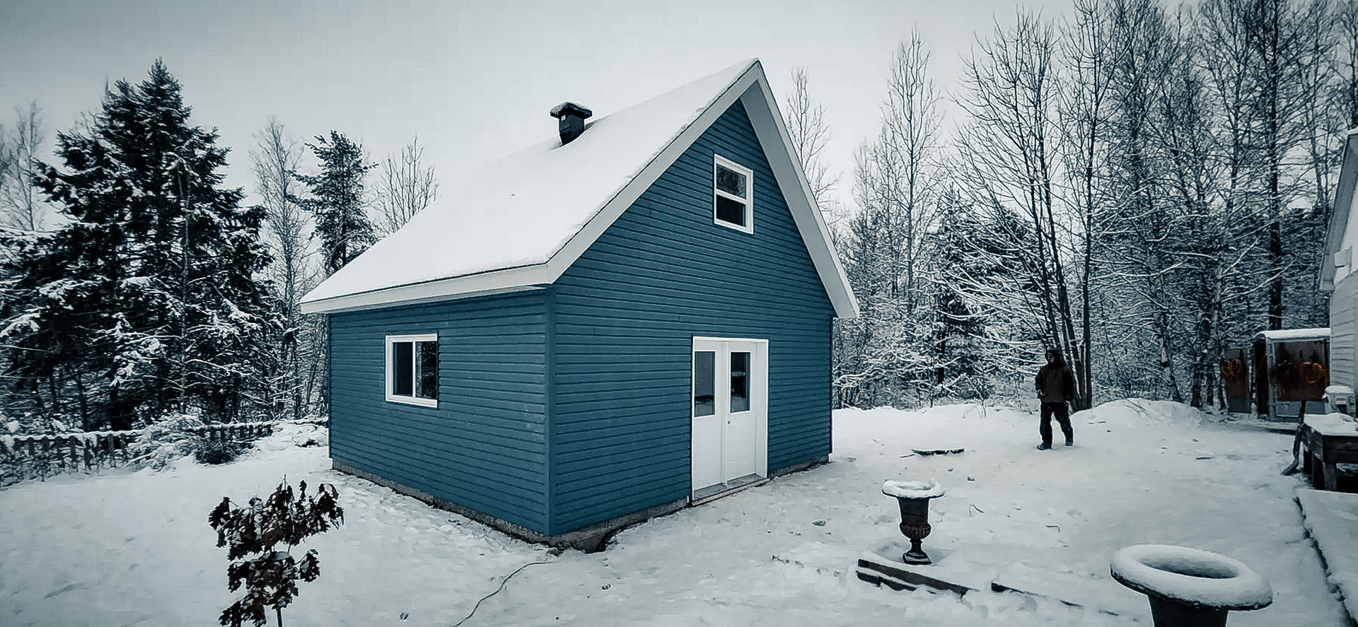 Small blue shed with a snow-covered roof, located in a winter forest. Peaceful setting with snow-covered trees and a person in the background.