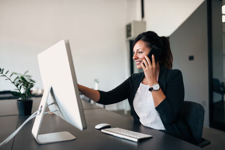 Smiling professional woman talking on the phone in front of a desktop computer in a modern workspace.