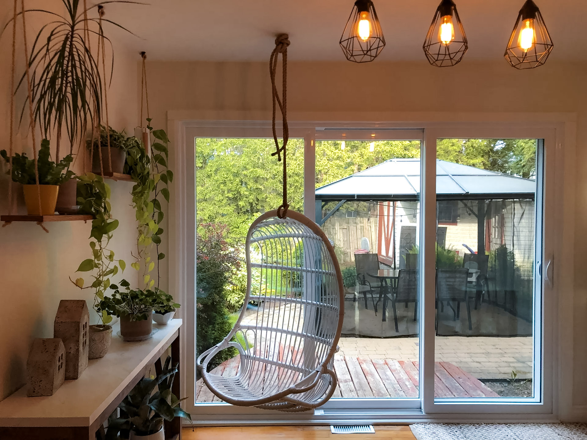 Coin détente intérieur avec chaise suspendue, grandes portes patio vitrées, plantes décoratives et vue sur terrasse avec gazebo.