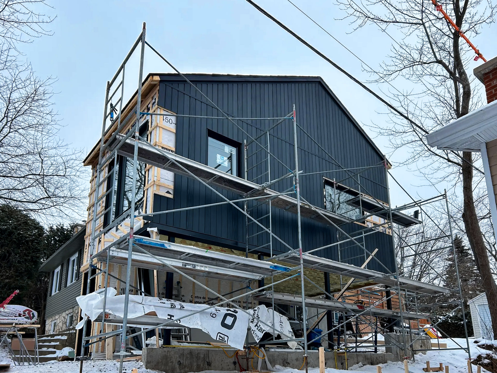 House under construction with scaffolding and installation of black vertical exterior cladding in winter