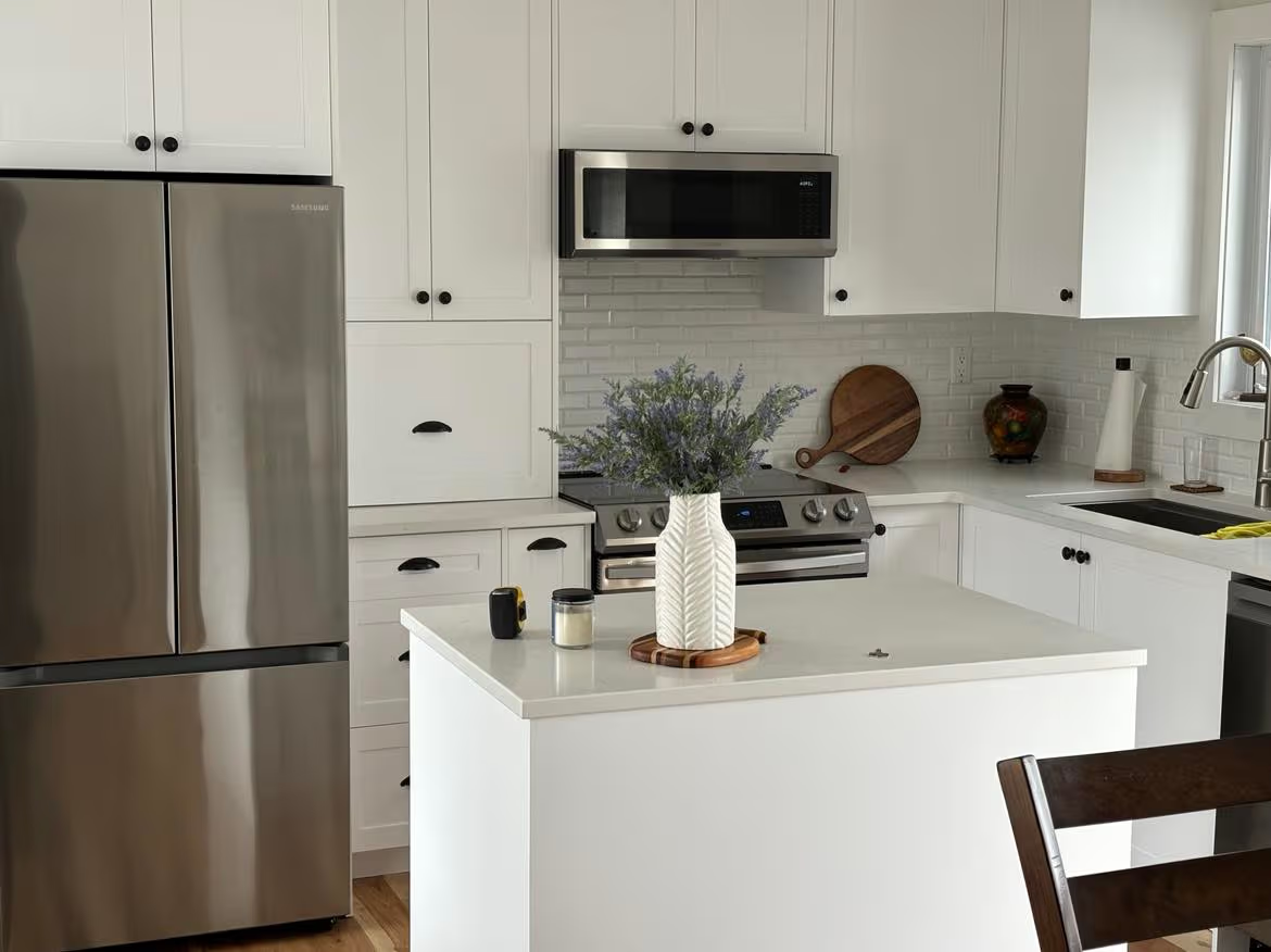 Cuisine blanche moderne avec îlot central, armoires shaker, électroménagers en inox, dosseret métro gris pâle, comptoir clair, évier sous fenêtre et décoration florale