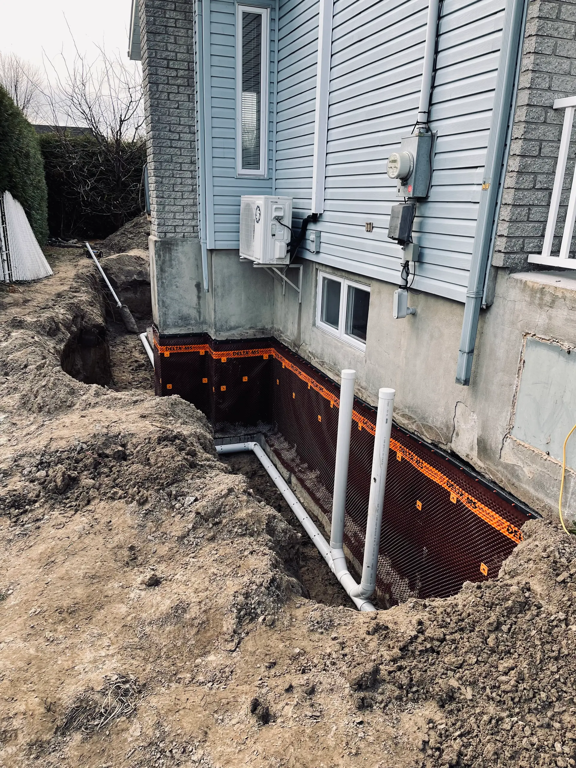 Travaux d'excavation pour l'installation d'un drain français et d'une membrane d'étanchéité sur la fondation d'une maison.