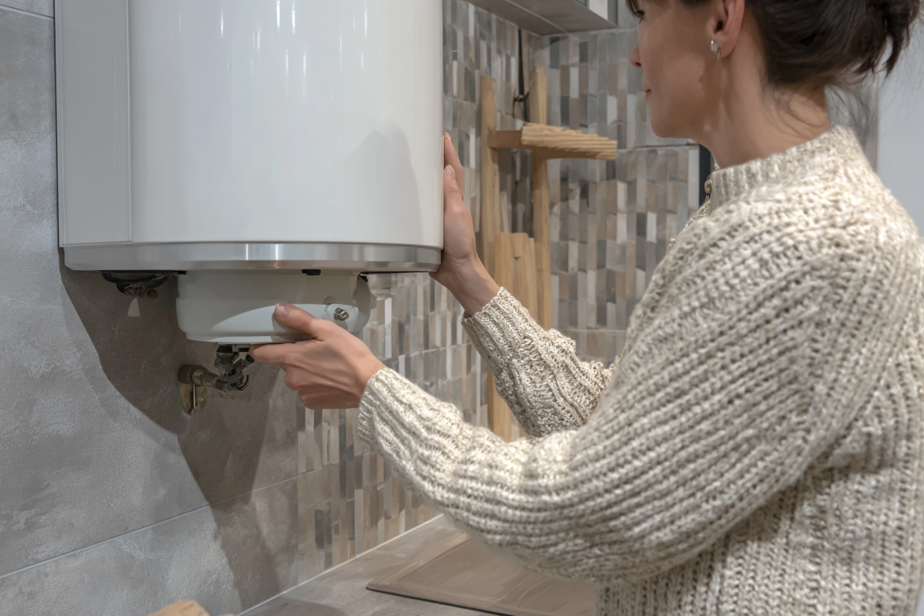 Woman checking a white wall-mounted water heater in a modern kitchen with ceramic tile backsplash