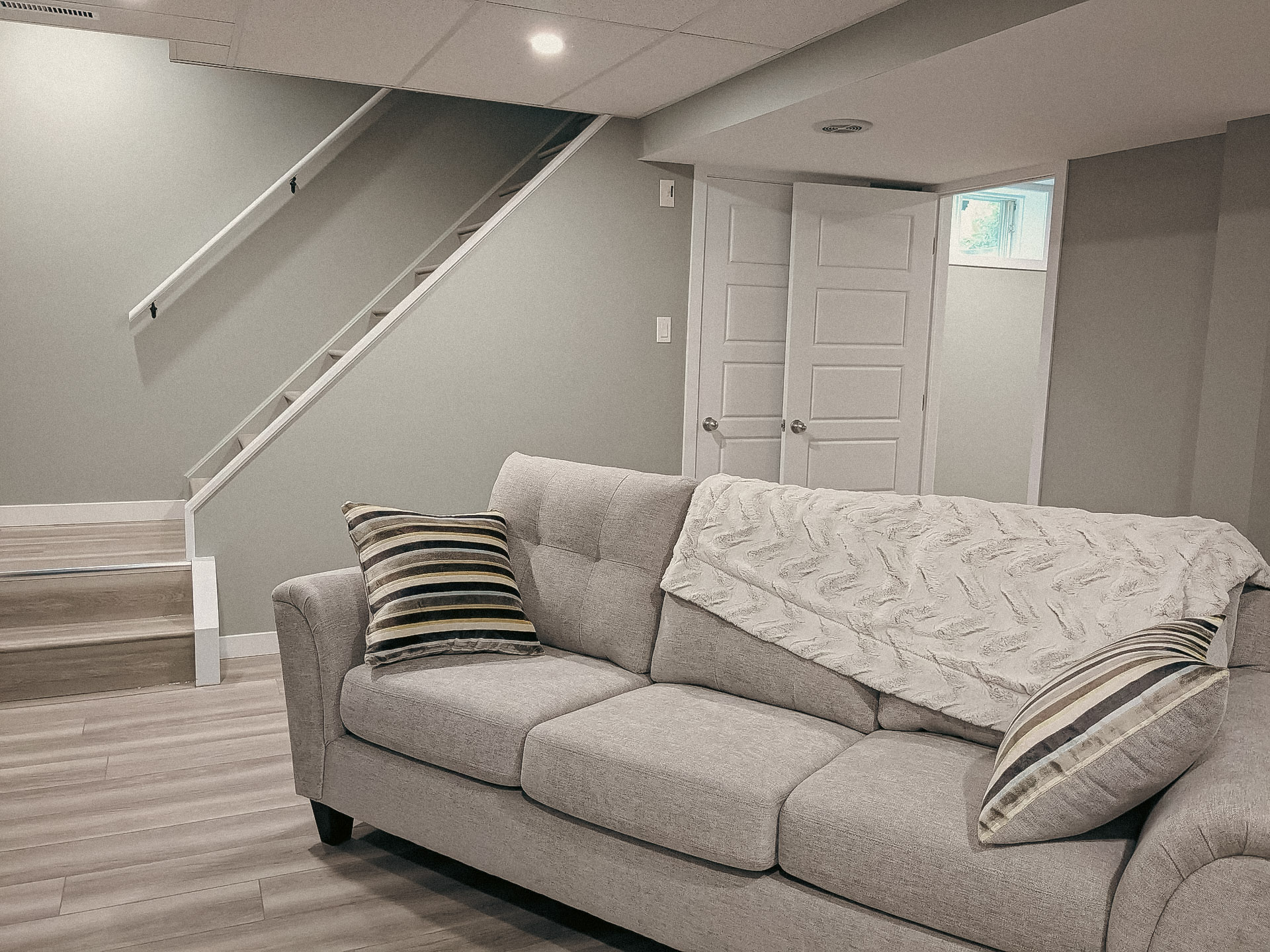 Sous-sol aménagé moderne avec canapé gris, plancher en bois, escalier ouvert, portes blanches et éclairage encastré.