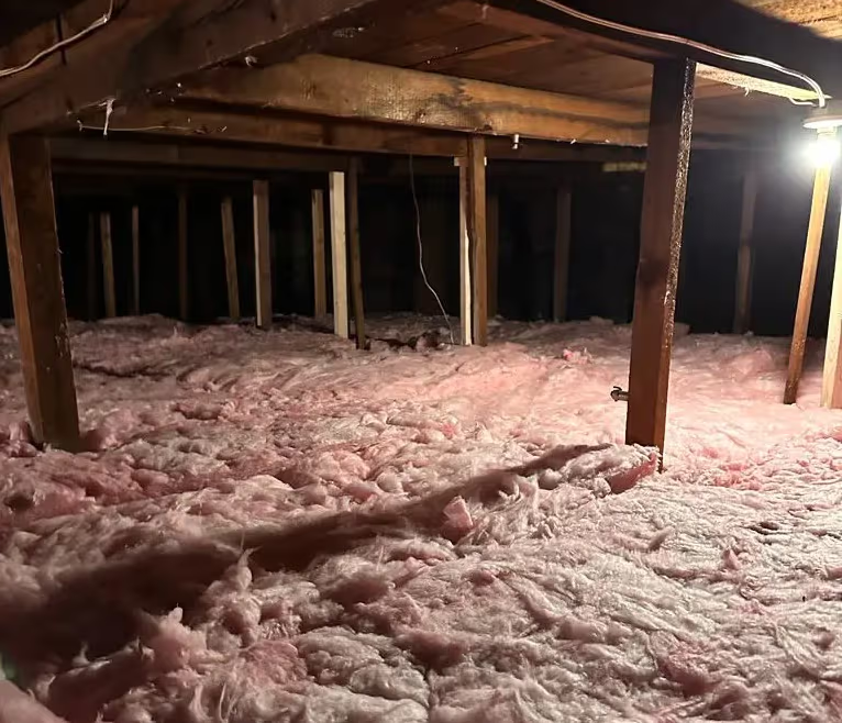 Insulated attic with pink blown-in insulation, wooden beams, exposed wiring and temporary lighting.