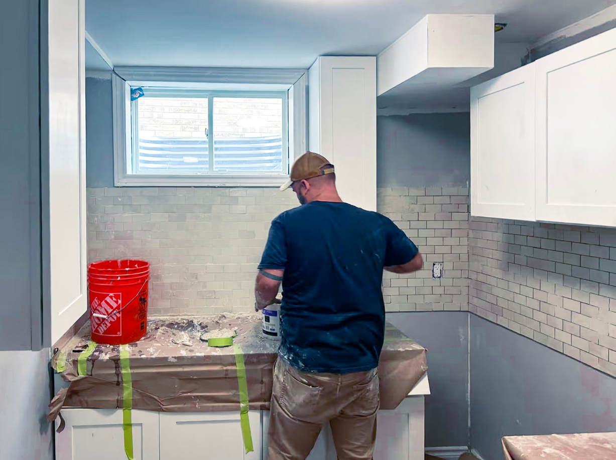 Travailleur appliquant du coulis sur un dosseret en céramique blanche dans une cuisine en rénovation avec comptoirs protégés.