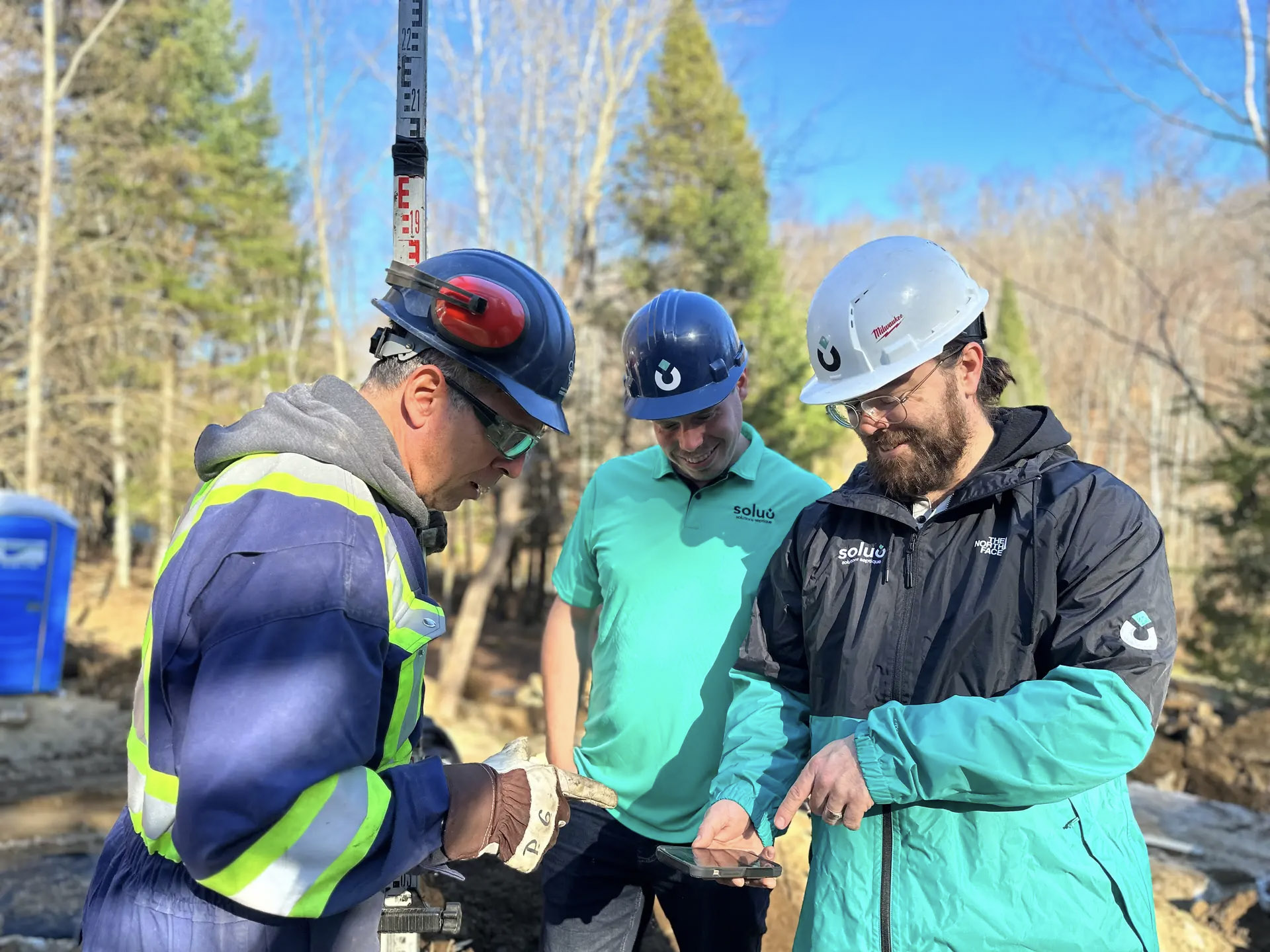 Three workers wearing safety helmets review information on a phone at an outdoor job site, illustrating teamwork and project coordination.