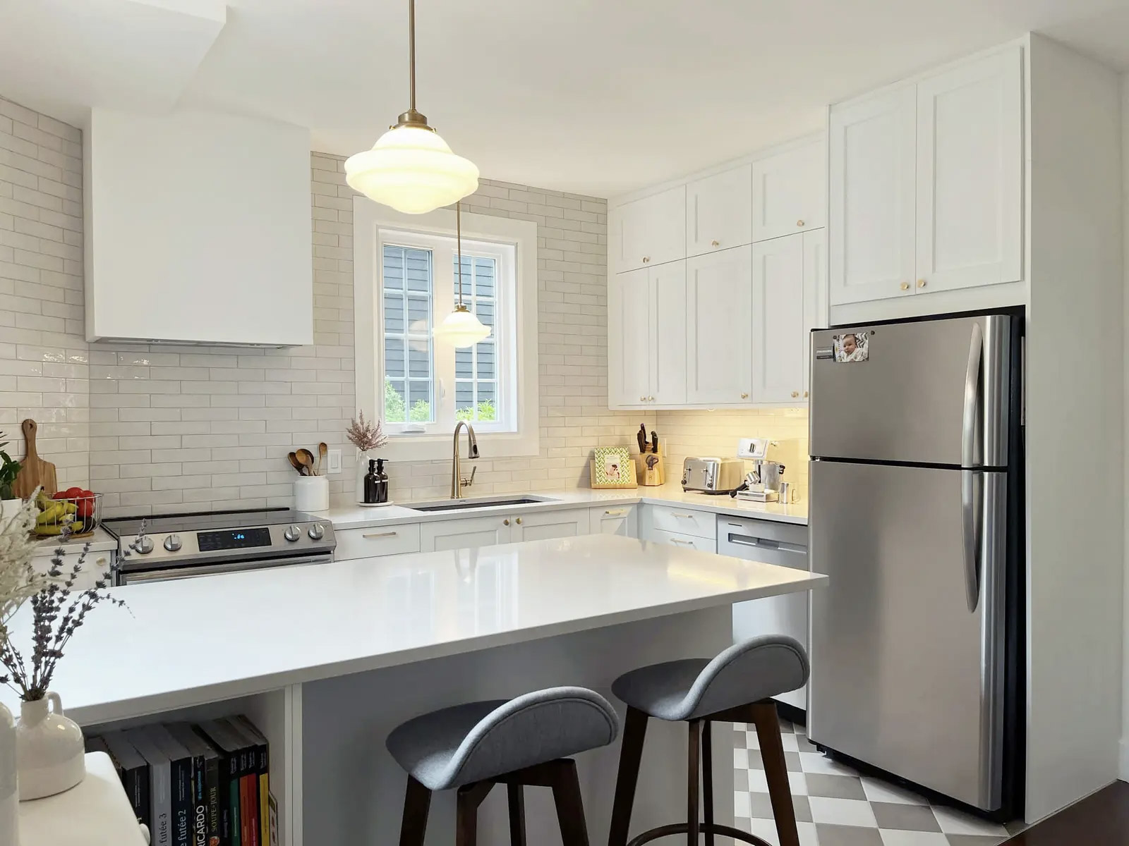 Cuisine blanche lumineuse avec grand îlot central, armoires pleine hauteur, comptoir blanc, dosseret en tuiles métro, réfrigérateur en acier inoxydable, tabourets gris, fenêtre et suspensions pour une ambiance moderne et accueillante.