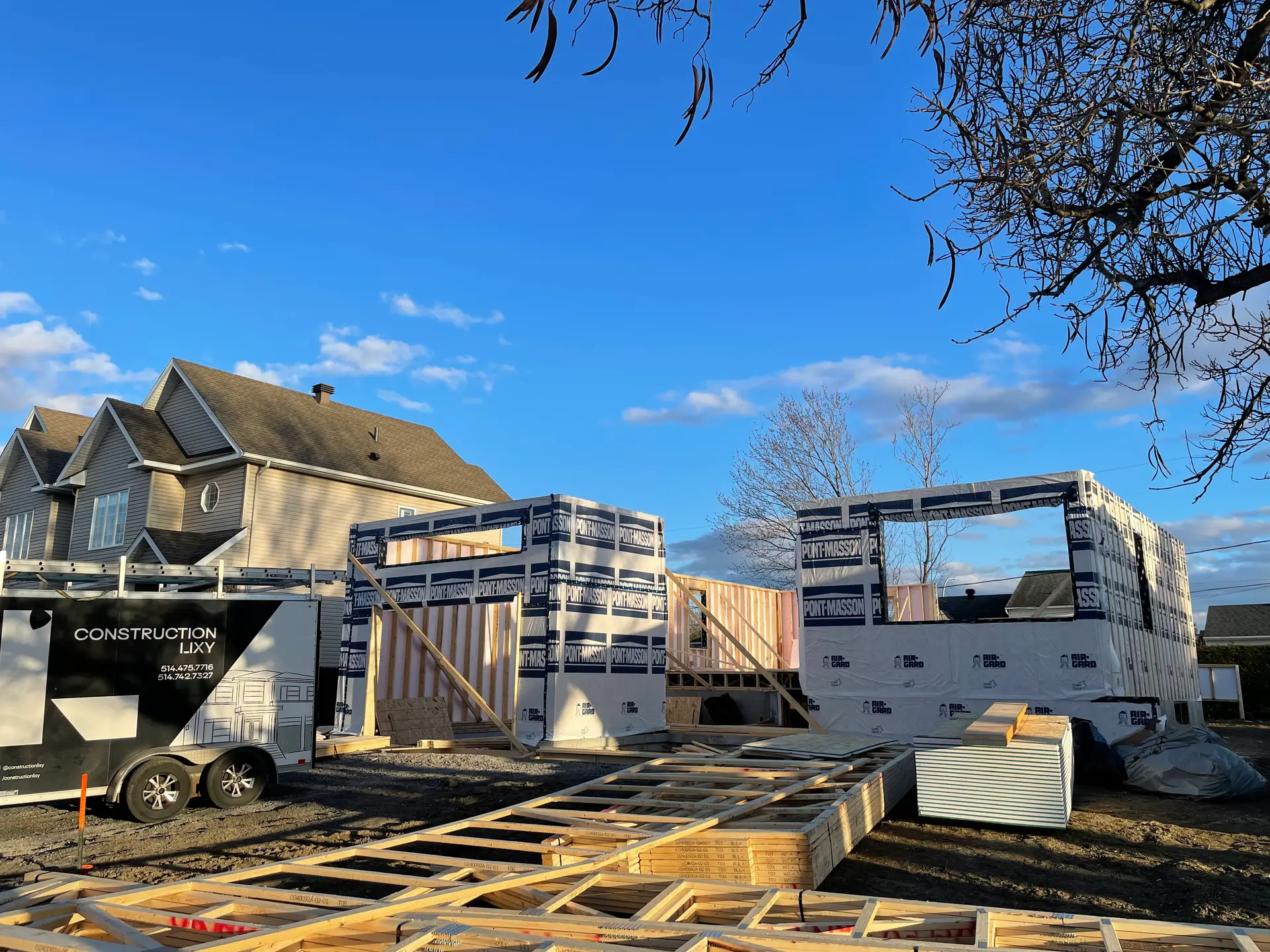 Construction d'une maison en ossature bois avec matériaux empilés et remorque de chantier au coucher du soleil.
