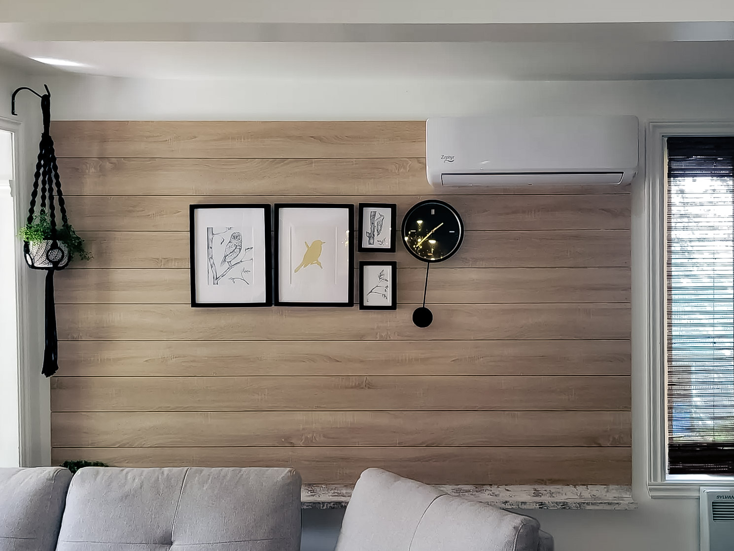 Light wood wall with wall-mounted air conditioning, decorative frame gallery, black designer clock and hanging plant in a modern living room.