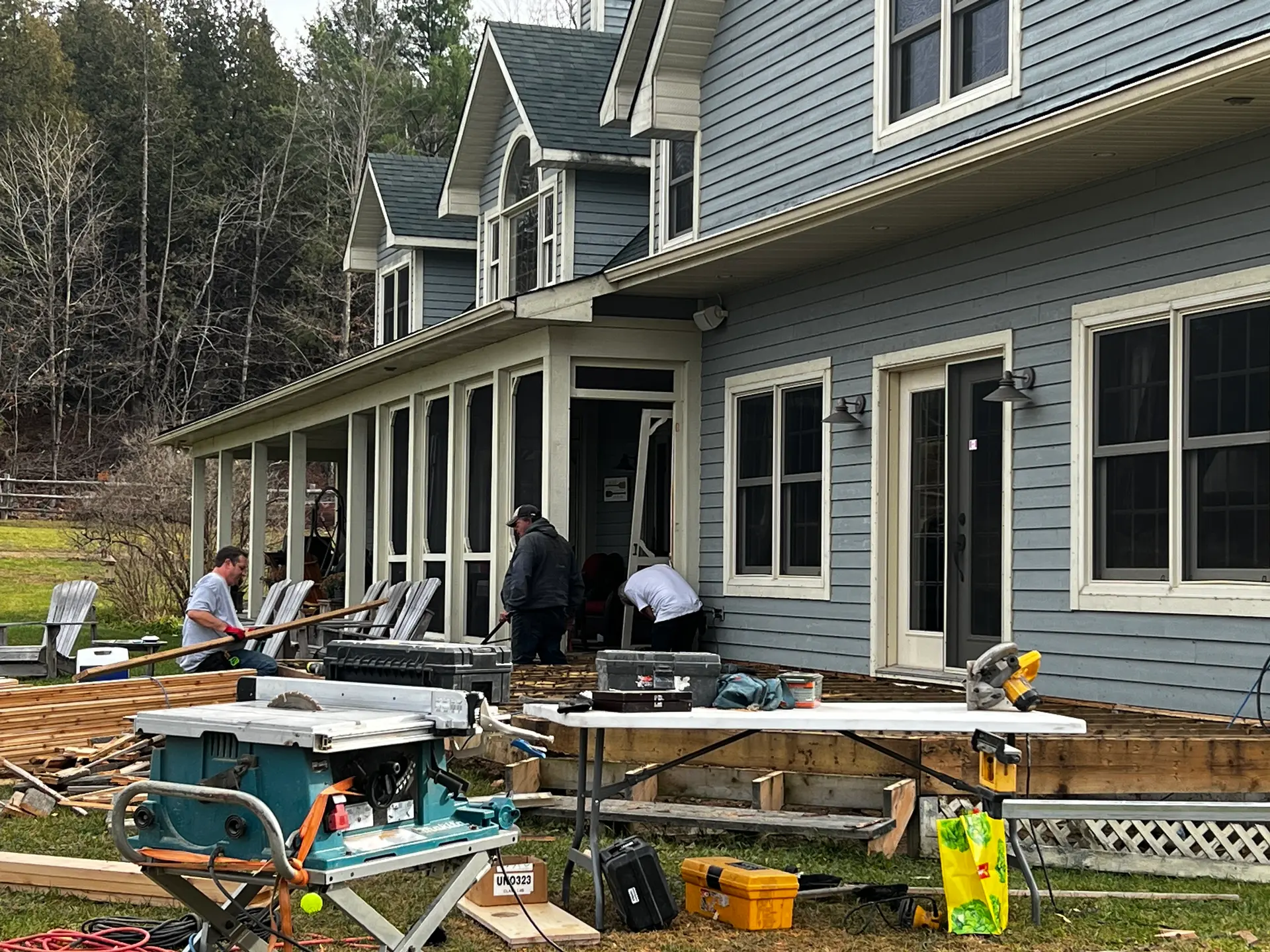 Workers renovating the wooden deck of a blue house with tools and construction equipment.